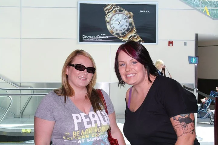 Two women standing in an indoor public space, smiling at the camera. One woman wears sunglasses and a gray t-shirt, the other has dark hair, a tattoo on her arm, and wears a black shirt.