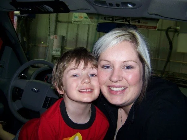 A woman and a young boy sitting together inside a vehicle, smiling at the camera.