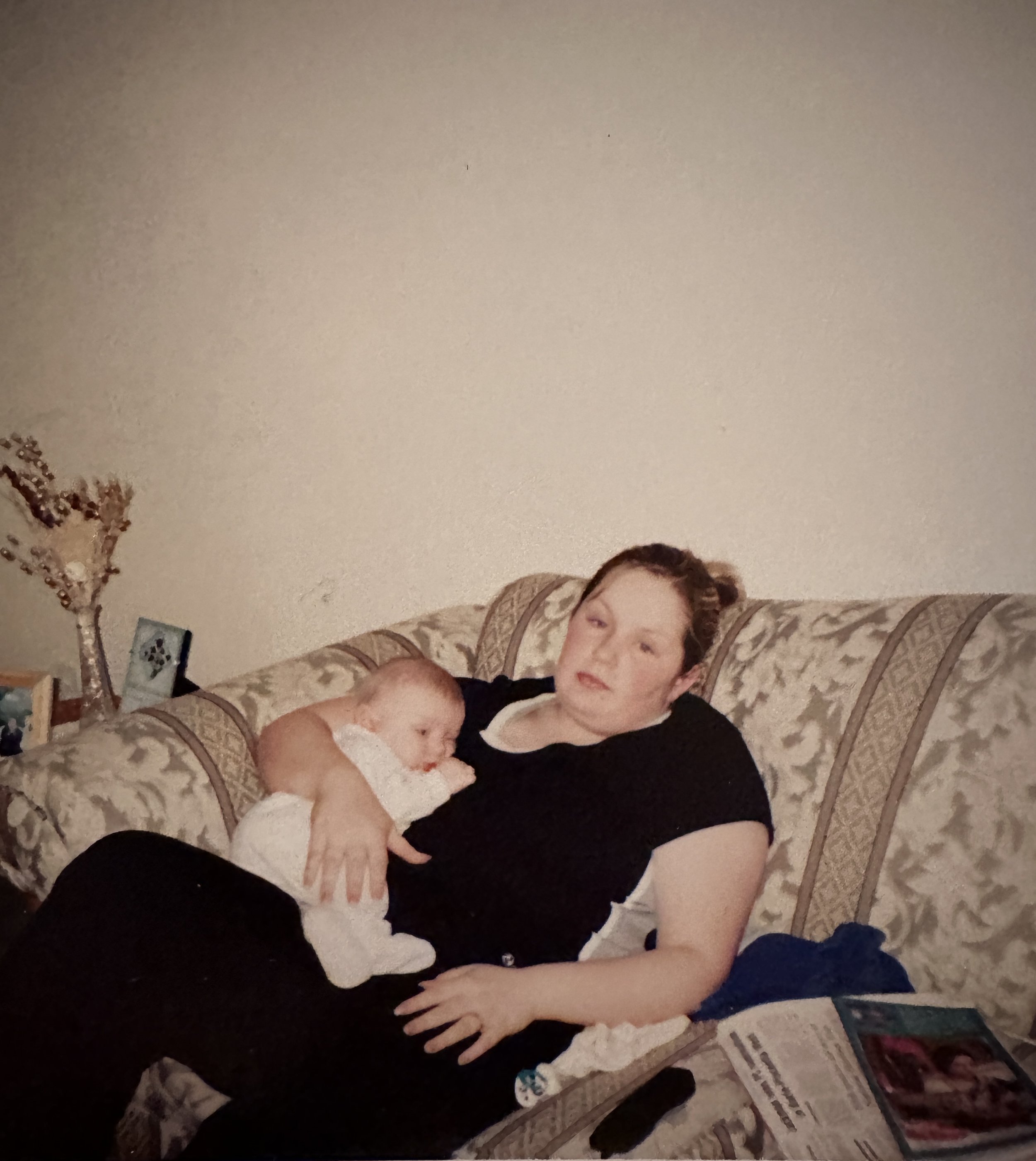 A woman sitting on a patterned sofa with a baby lying on her lap, the woman appears to be resting or sleeping, and the baby is looking to the side. There is a coffee table with various items, including magazines and remote controls, in front of the s