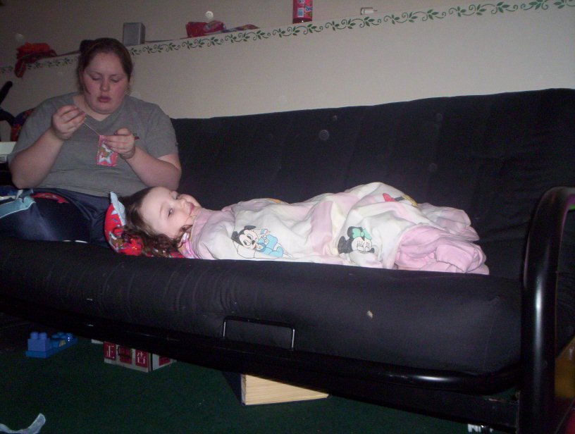 Child lying on a black couch wrapped in a Disney-themed blanket, with a woman sitting next to her on the couch, looking at something in her hands.