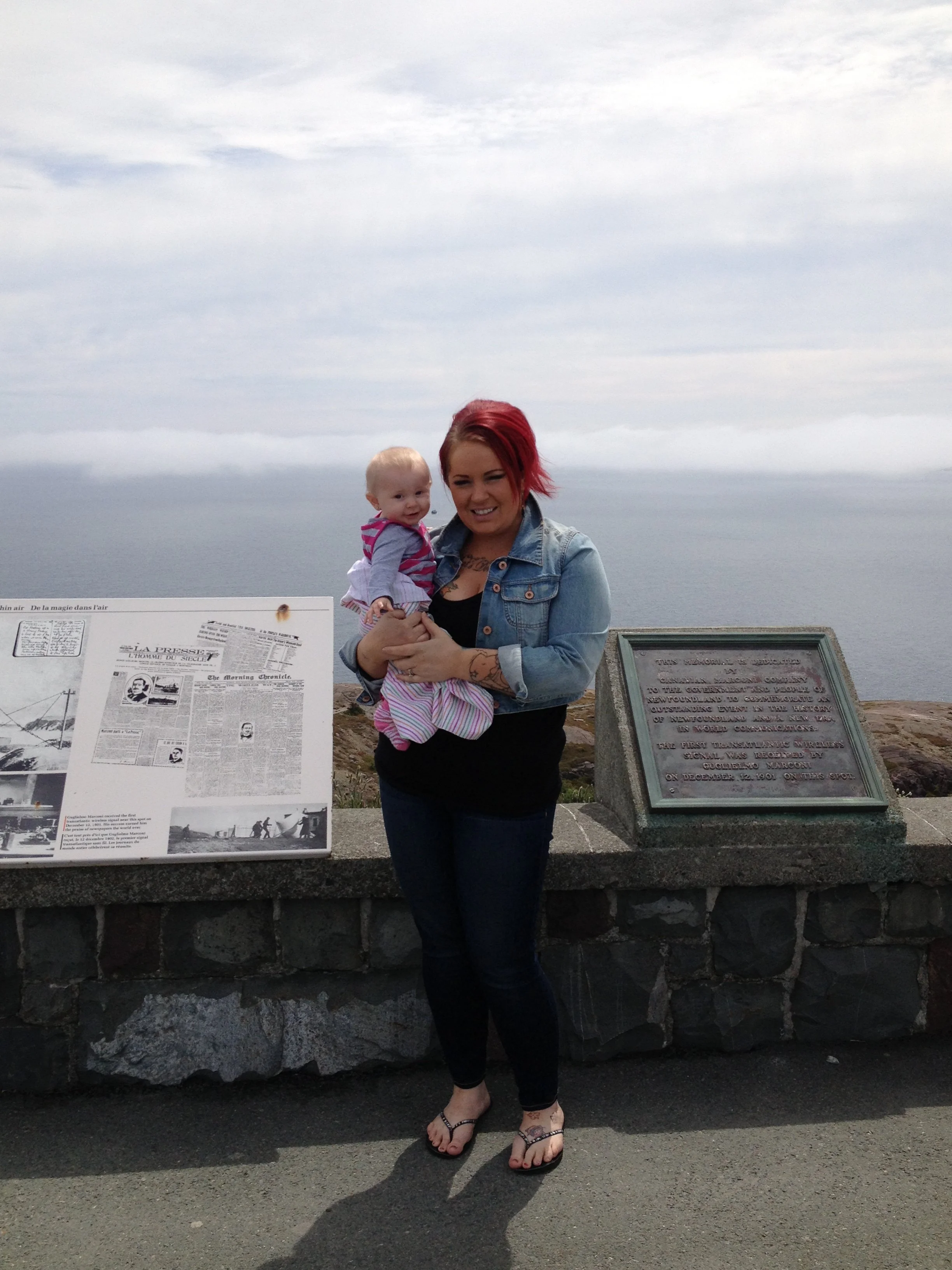 A woman with red hair holding a toddler girl with blonde hair, standing outdoors near a stone ledge with informational plaques, overlooking the water and cloudy sky.