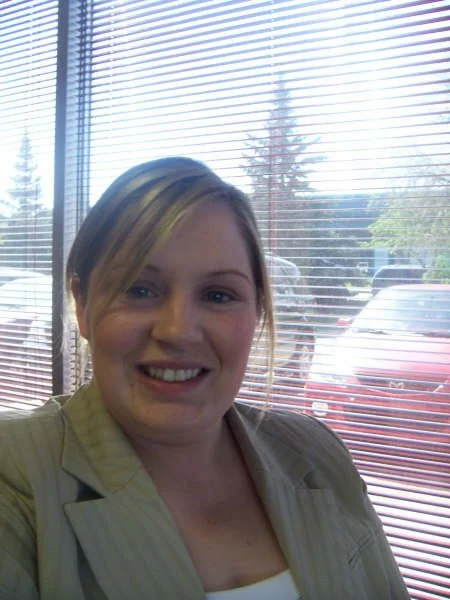 A woman taking a selfie inside near a window with blinds. Outside, parked cars and trees are visible.