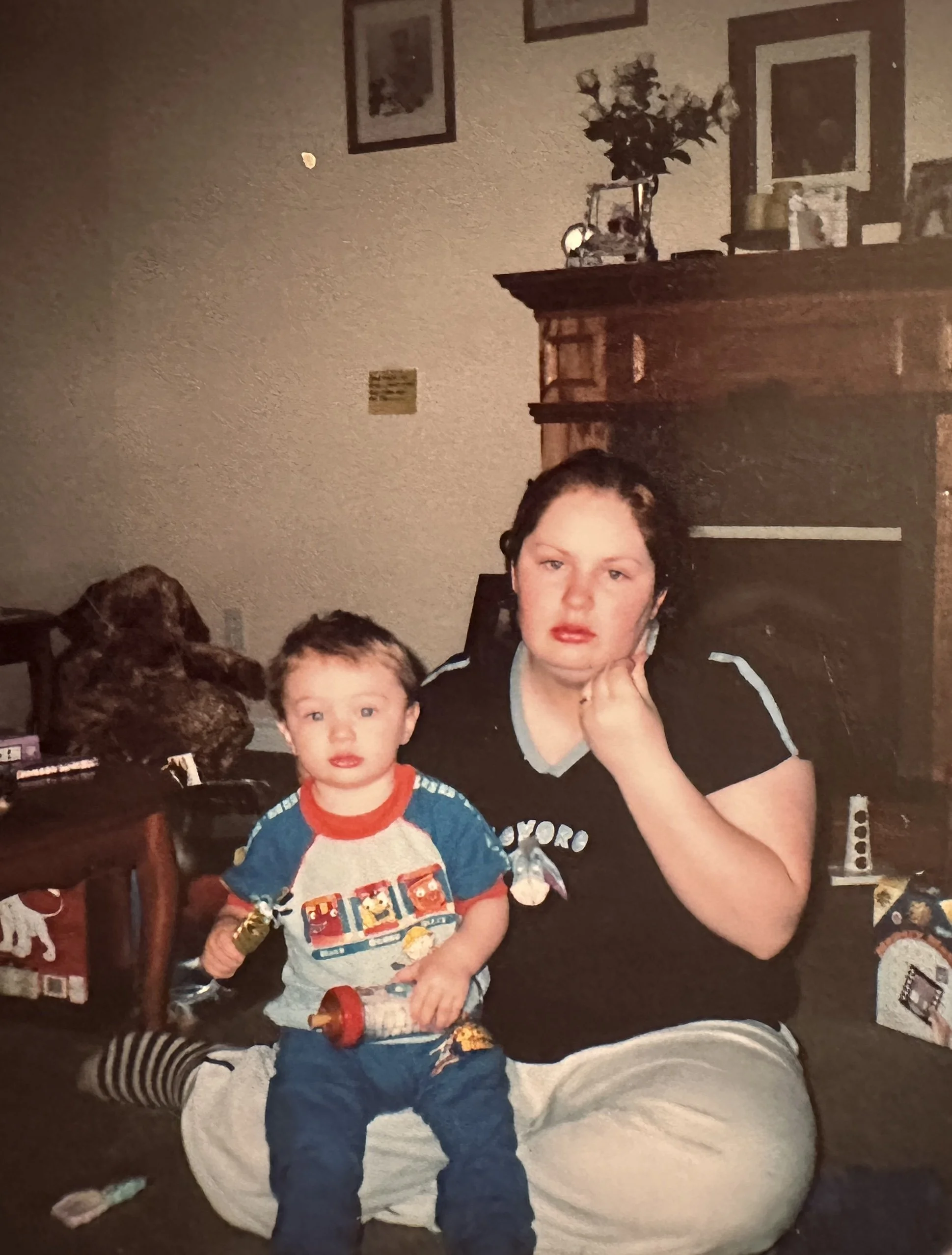 A woman with short dark hair and a black shirt sitting on the floor with her chin resting on her hand, next to a young boy with short dark hair holding a toy and looking at the camera, in a living room with a wooden fireplace, picture frames, and hol