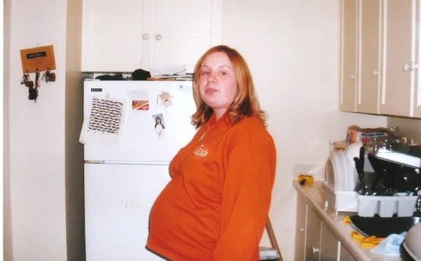 A woman with red hair in a kitchen, wearing an orange shirt, standing near a white refrigerator.
