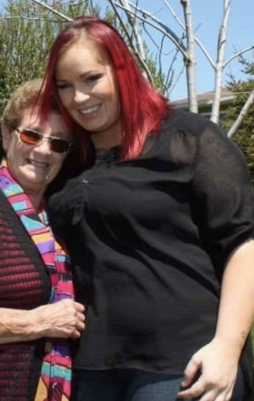 Two women smiling outdoors, one with short hair and sunglasses, the other with long red hair, posing together.