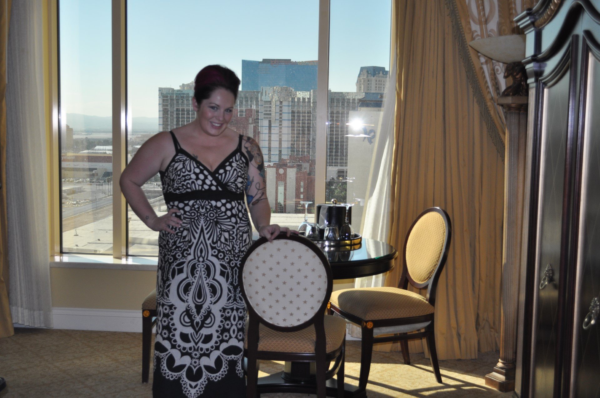 Woman in a black and white patterned dress standing next to a small round table with a tray, teapot, and cups, inside a room with large windows and city buildings in the background.