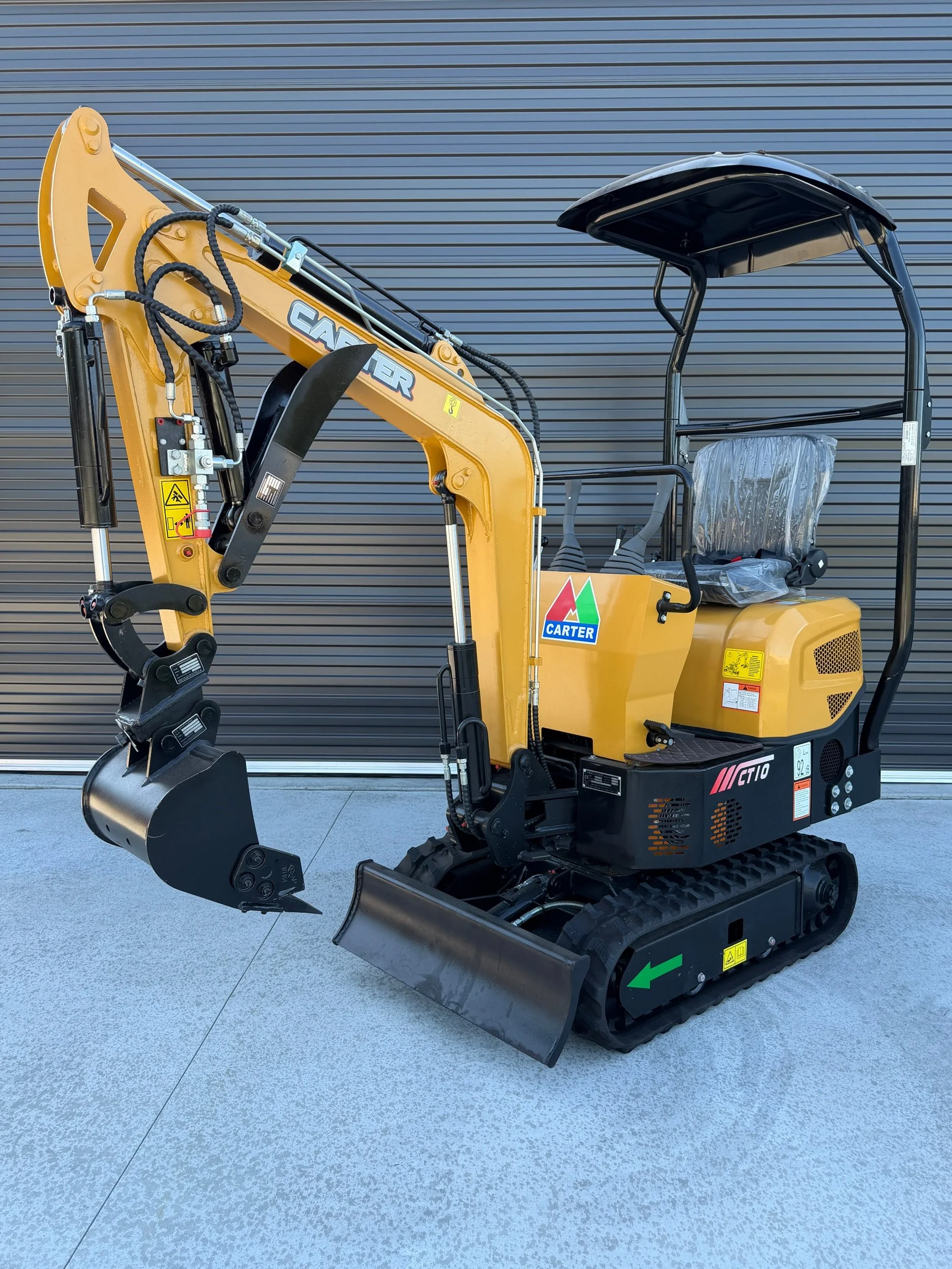 Yellow compact excavator with black tracks, black arm and bucket, and a gray canopy, parked on a concrete surface in front of a closed, horizontal metal door.