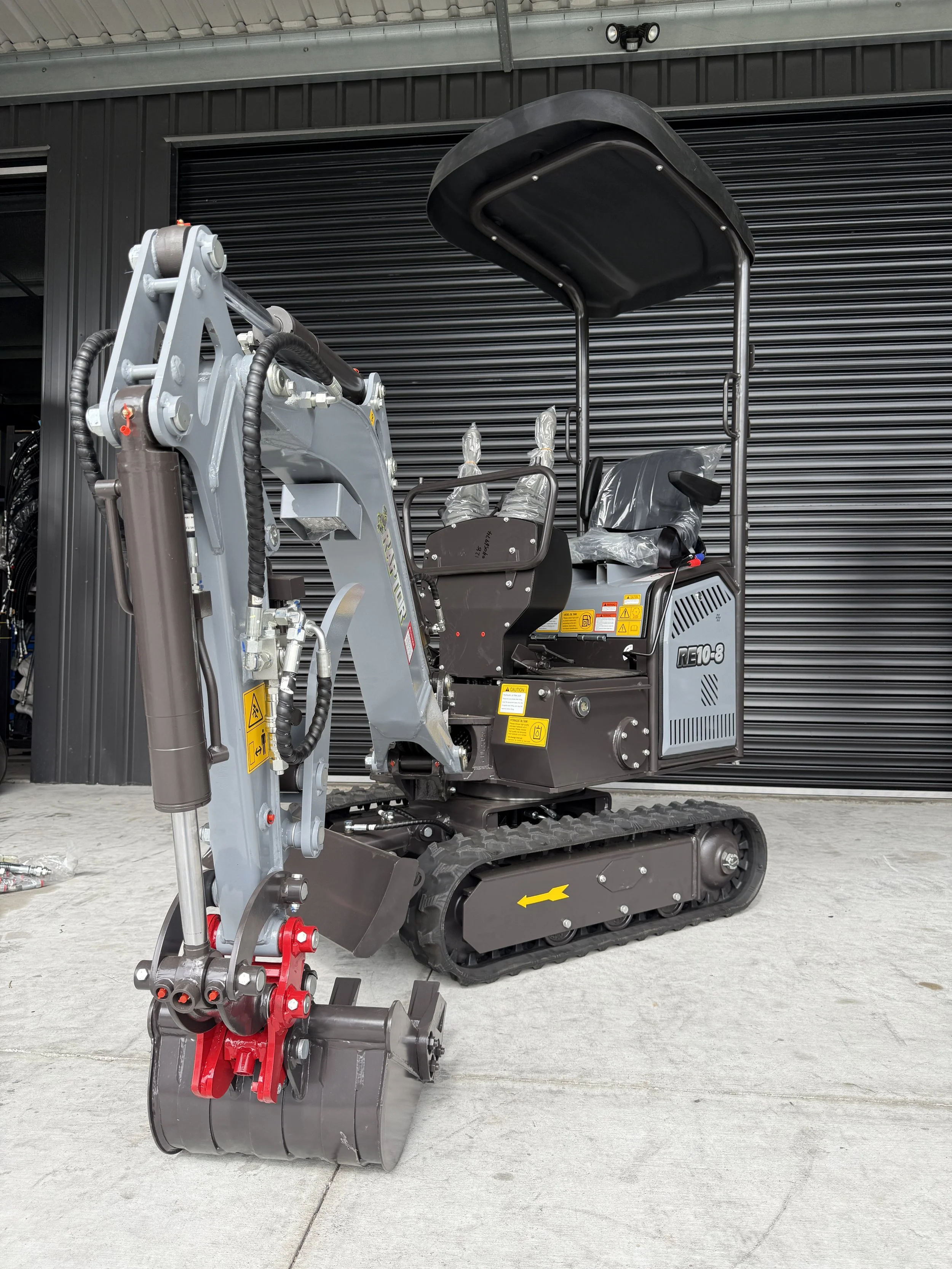 Small tracked excavator with a black canopy, grey and black body, and red hydraulic components, positioned in front of a black metal roller shutter door.