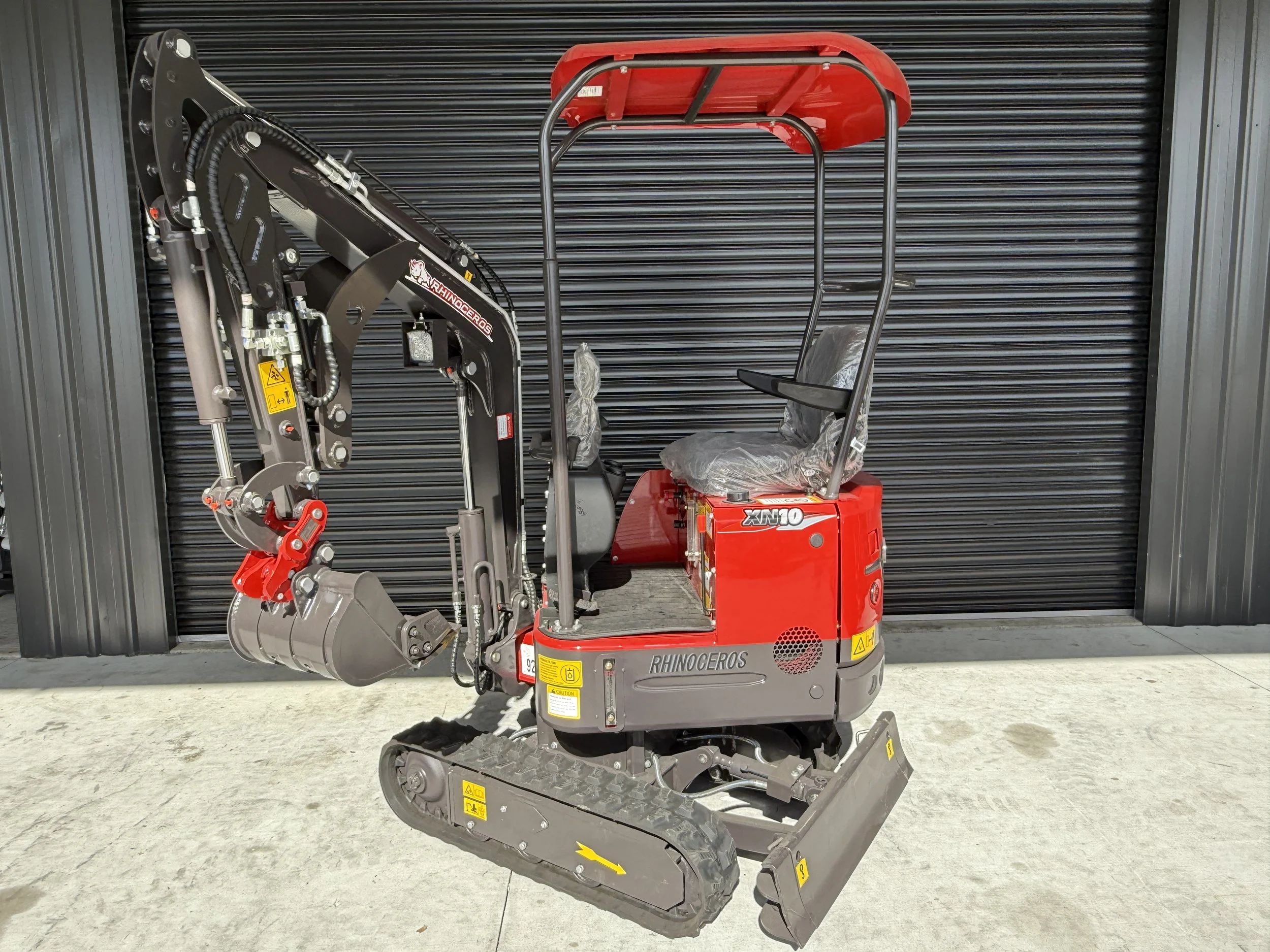 Small red mini excavator with tracks, parked in front of black metal garage door.