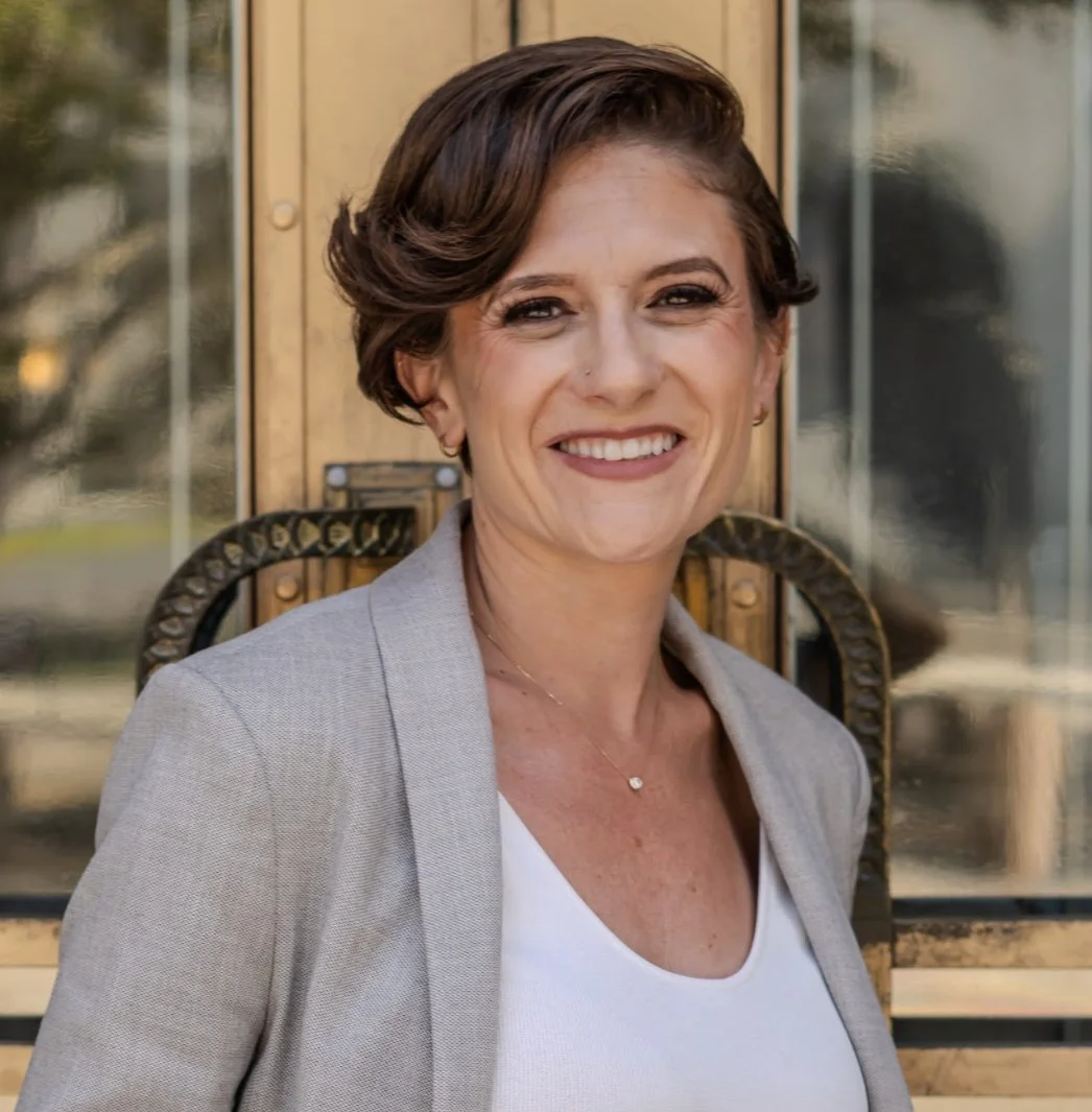 A woman with short brown hair, wearing a light grey blazer over a white top, navy blue pants with gold buttons, standing outdoors in front of a glass door with a reflection of trees.