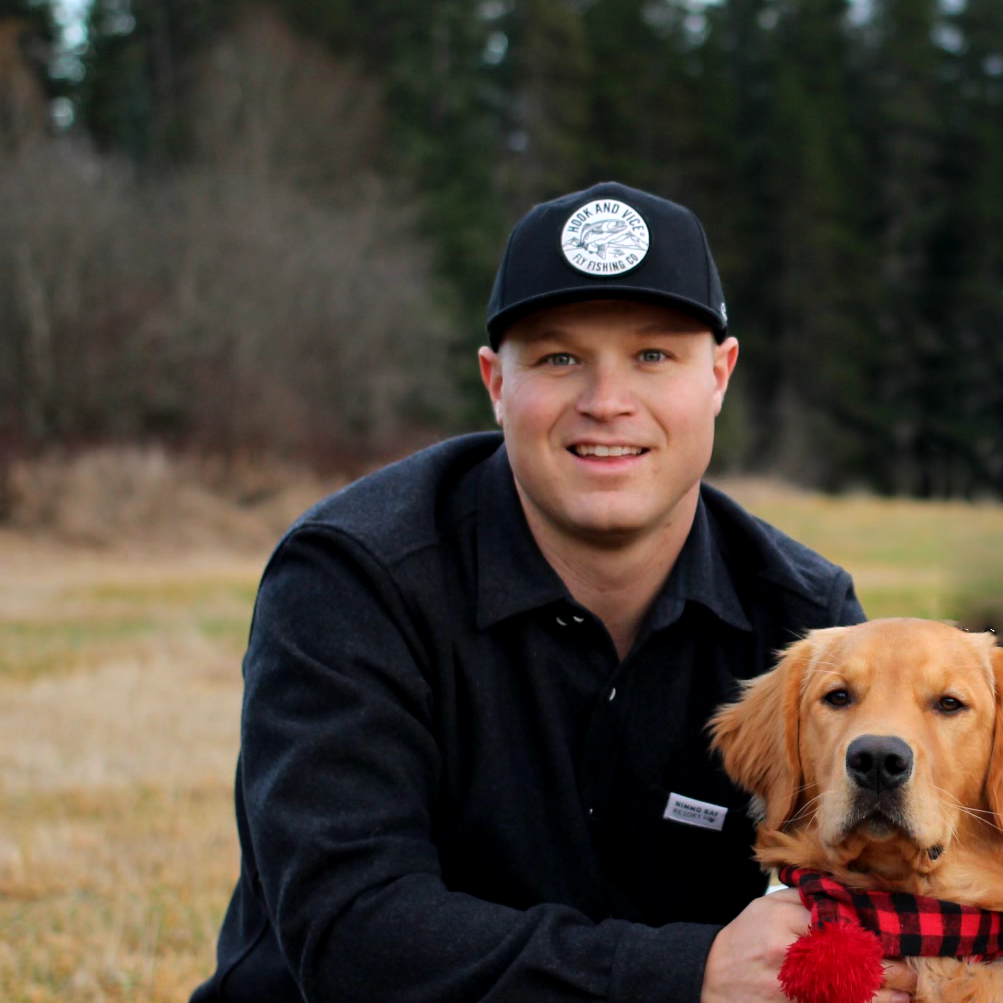 Outdoor portrait of a quality consulting professional wearing a black cap and jacket, kneeling in a field with a golden retriever.