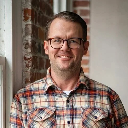 Professional portrait of a quality management consultant wearing glasses and a plaid shirt, standing by a window in an industrial-style office.