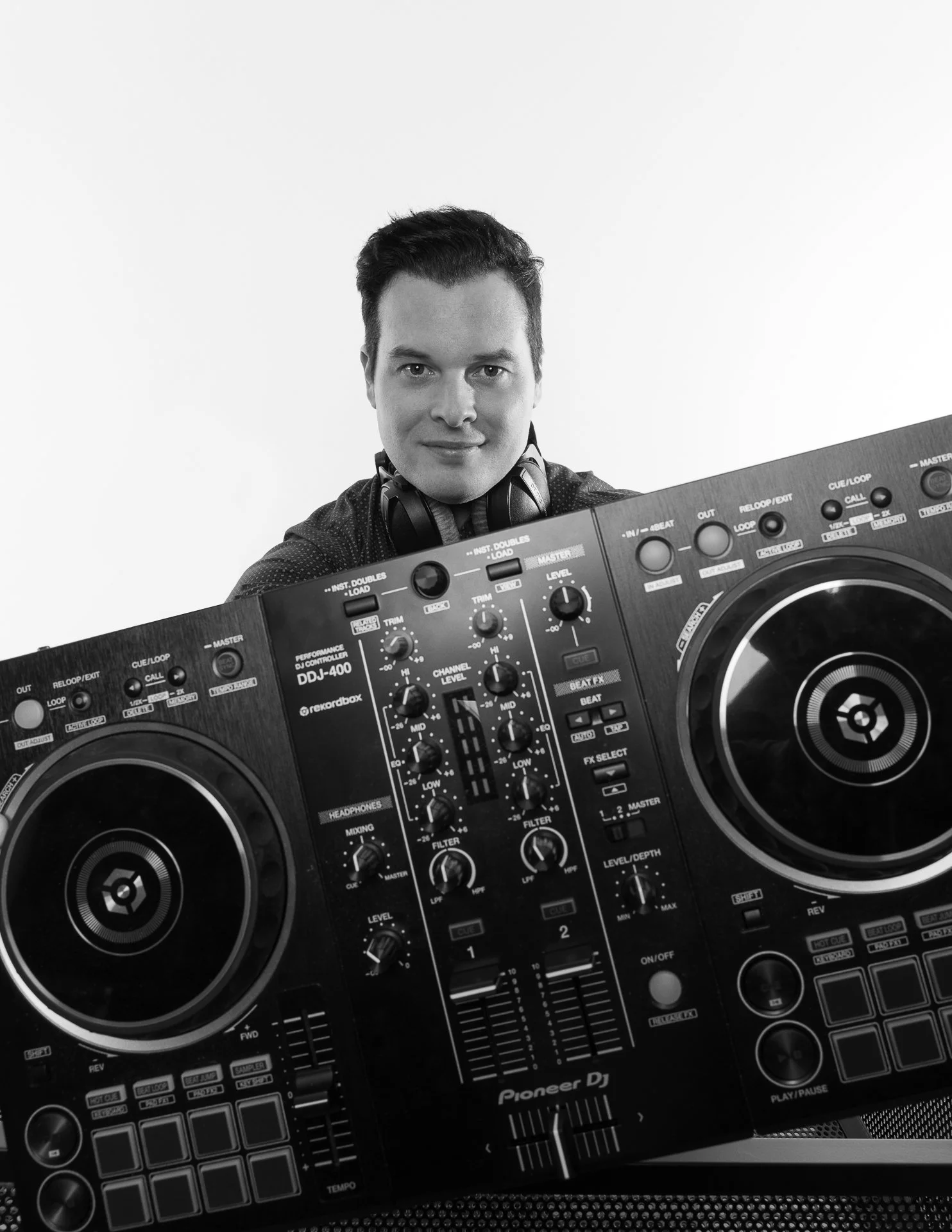 A man with dark hair wearing headphones around his neck standing behind DJ equipment, including a Pioneer DJ controller with various knobs, buttons, and two large jog wheels.