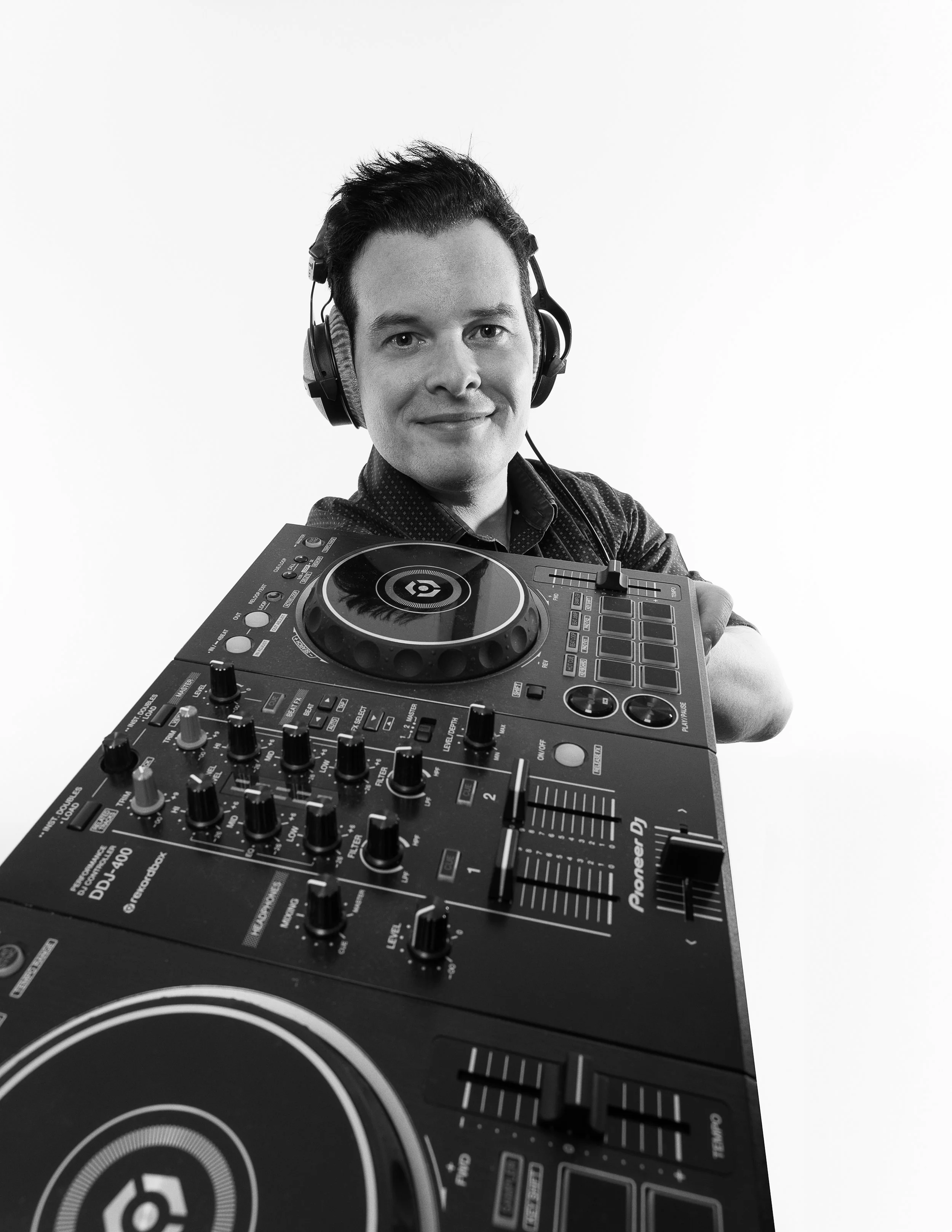 Black and white photo of a male DJ standing behind his DJ equipment, wearing headphones and looking at the camera.