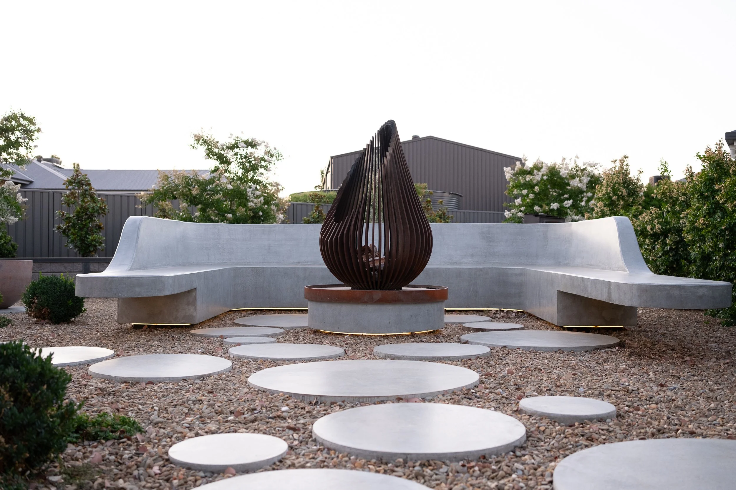 Outdoor seating area with curved white concrete benches, a central decorative sculpture made of vertical wooden slats, stepping stones on gravel, greenery including bushes and flowering trees, and buildings with metal siding in the background.