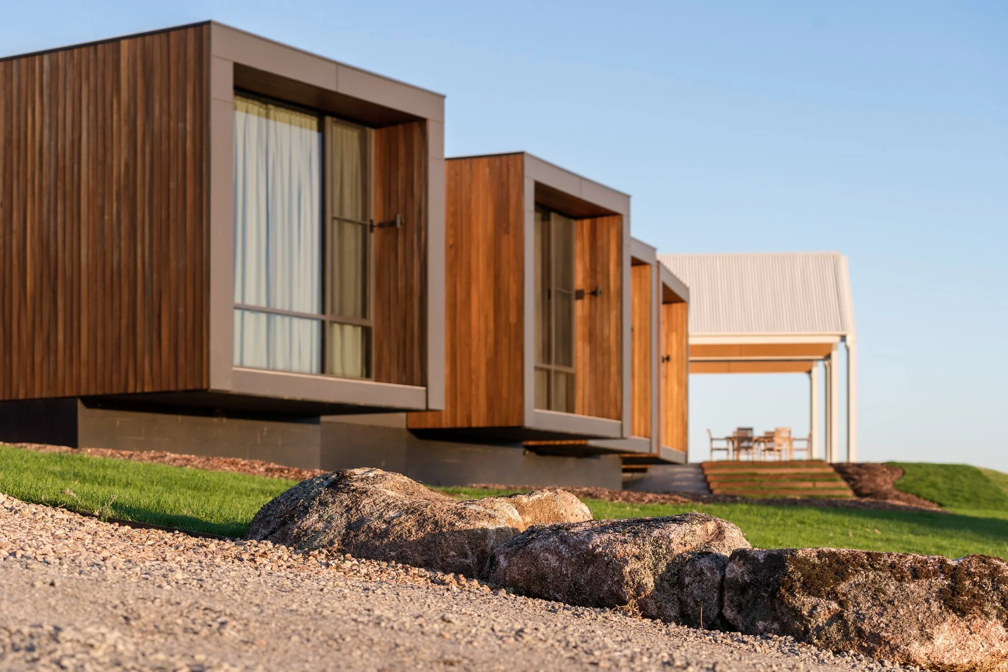Modern house with wooden and white exterior, large windows, and outdoor patio, under clear blue sky, with rocks and grass in foreground.