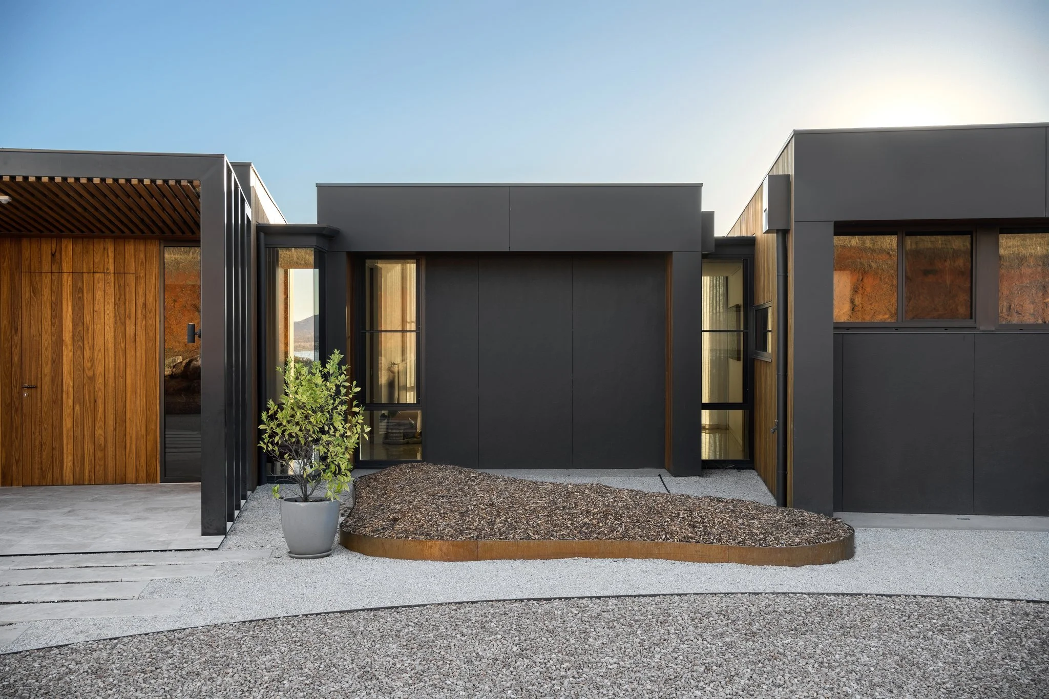 Modern house with dark gray and wood exterior, large windows, and landscaped front yard with gravel and a small potted shrub.