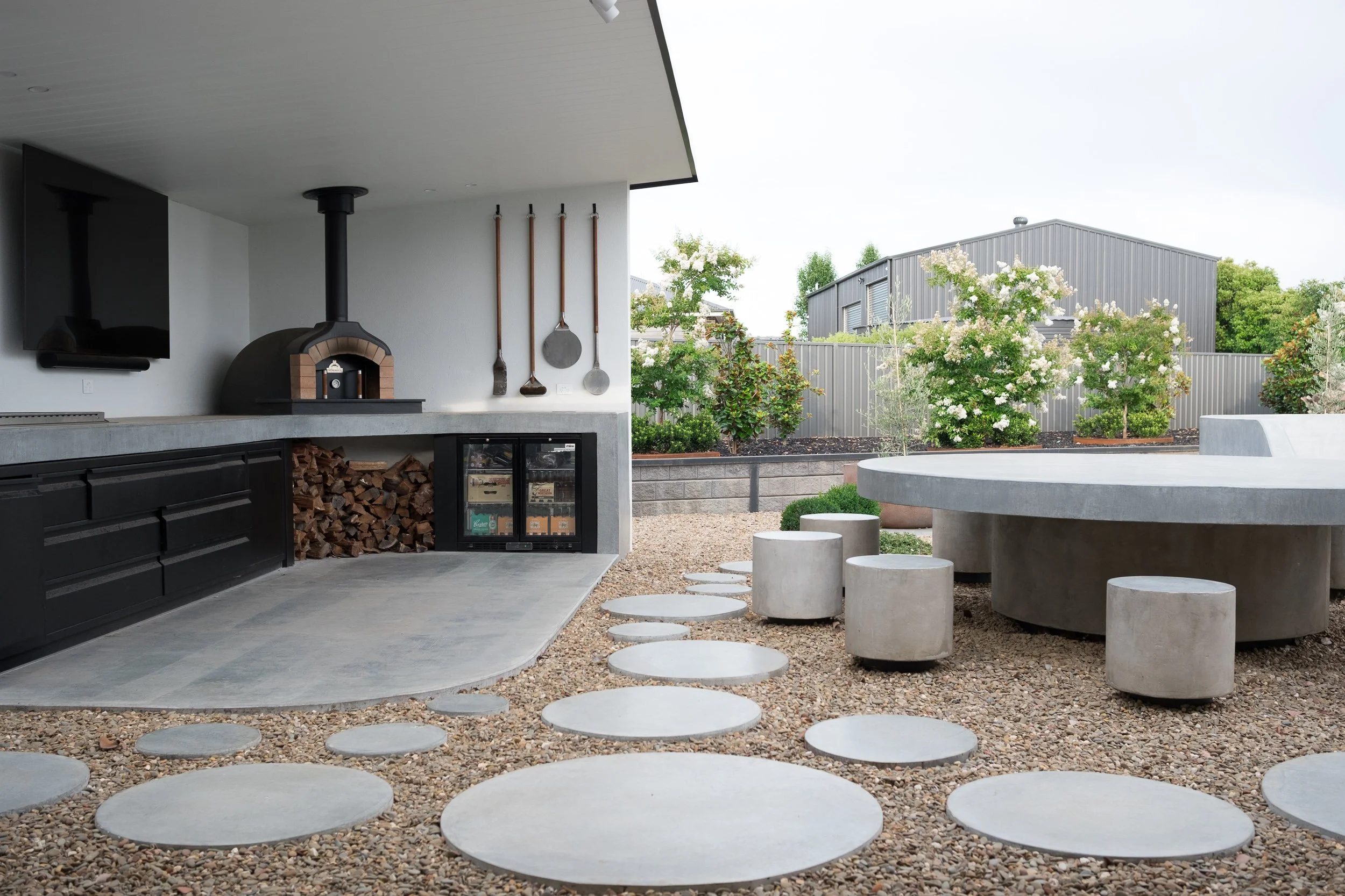Modern outdoor patio area with concrete circular stepping stones on gravel, a built-in outdoor kitchen with wood-fired oven, and lush greenery including flowering bushes and trees.