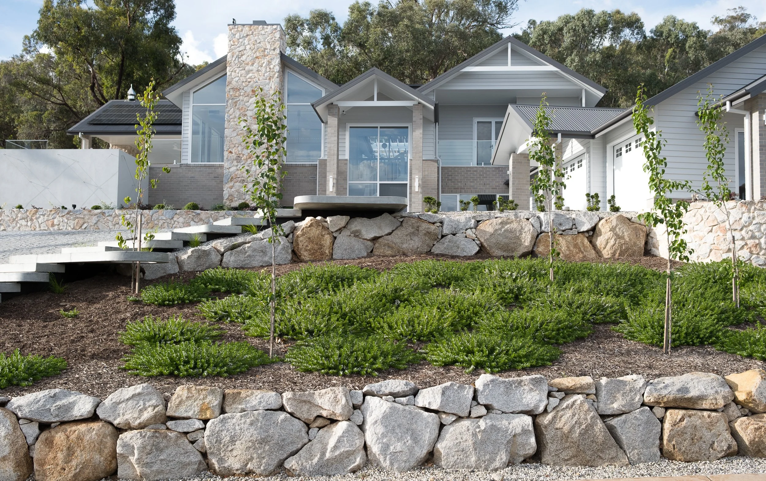 Modern house with large glass windows, stone chimney, and solar panels on the roof, surrounded by landscaped garden with young trees and rocks.