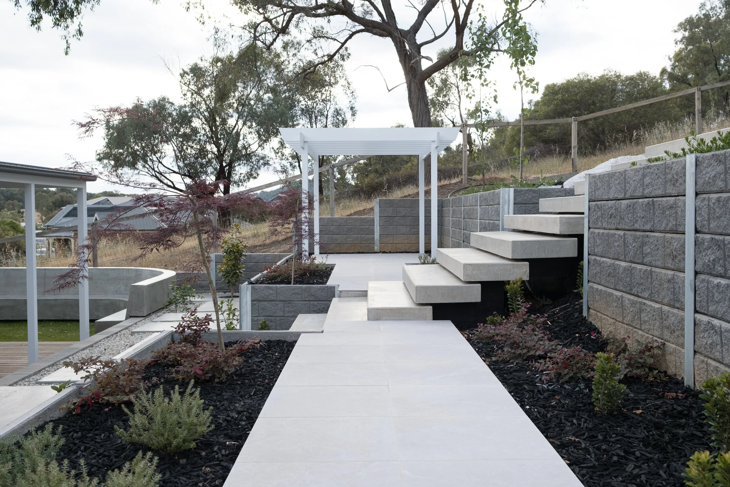 A backyard with modern landscaping, including concrete stairs, a white pergola, and various plants and trees. There is a stone retaining wall and a paved pathway.