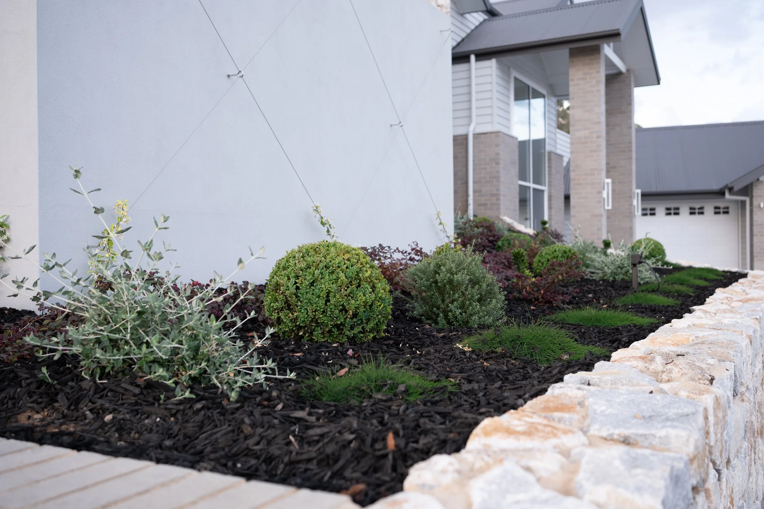 A landscaped front yard with a garden bed filled with various shrubs and plants surrounded by dark mulch, bordered by a row of light-colored rocks, with a modern house featuring brick and siding in the background.