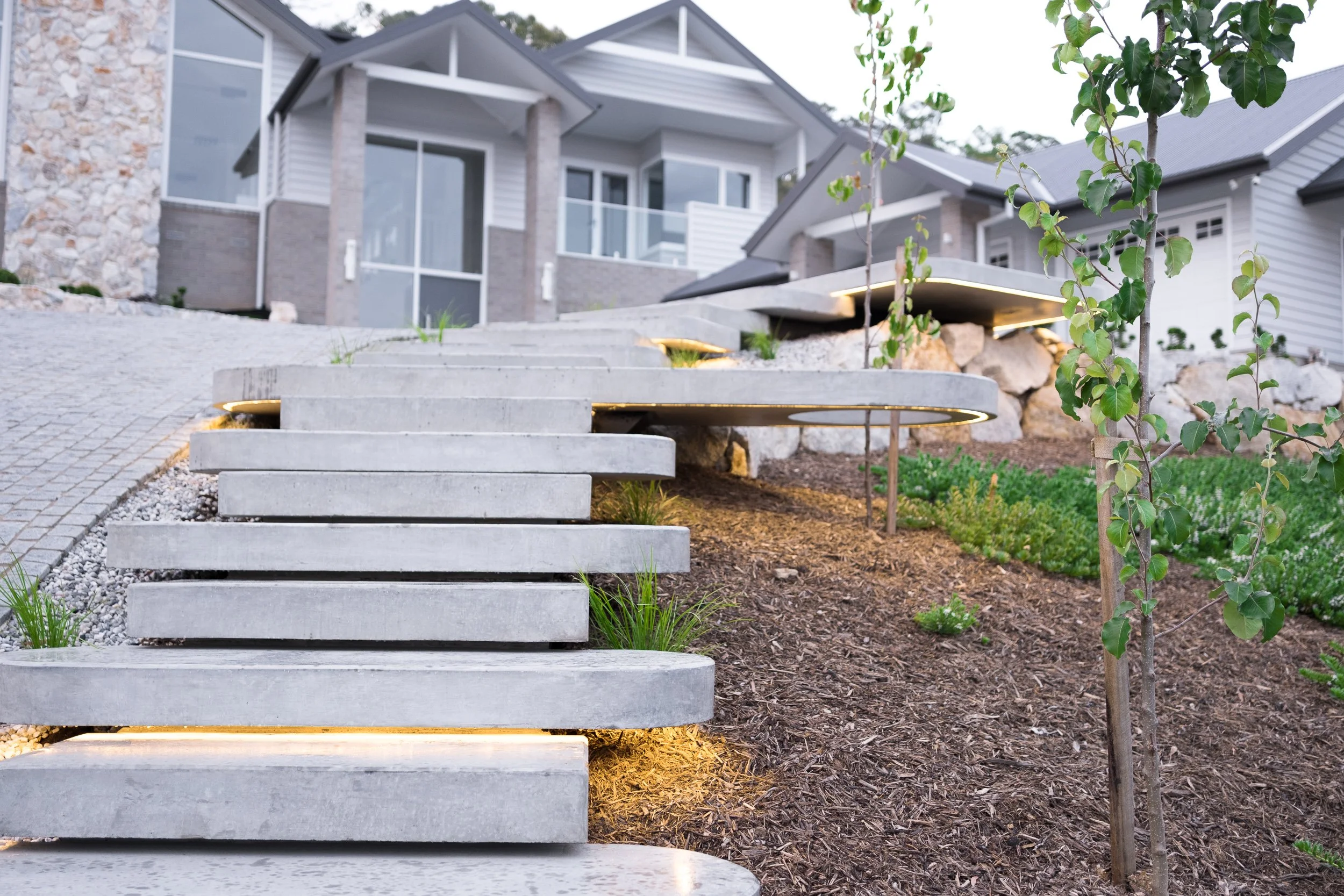Concrete stairs leading up to a modern house with large windows, surrounded by landscaped plants and trees.