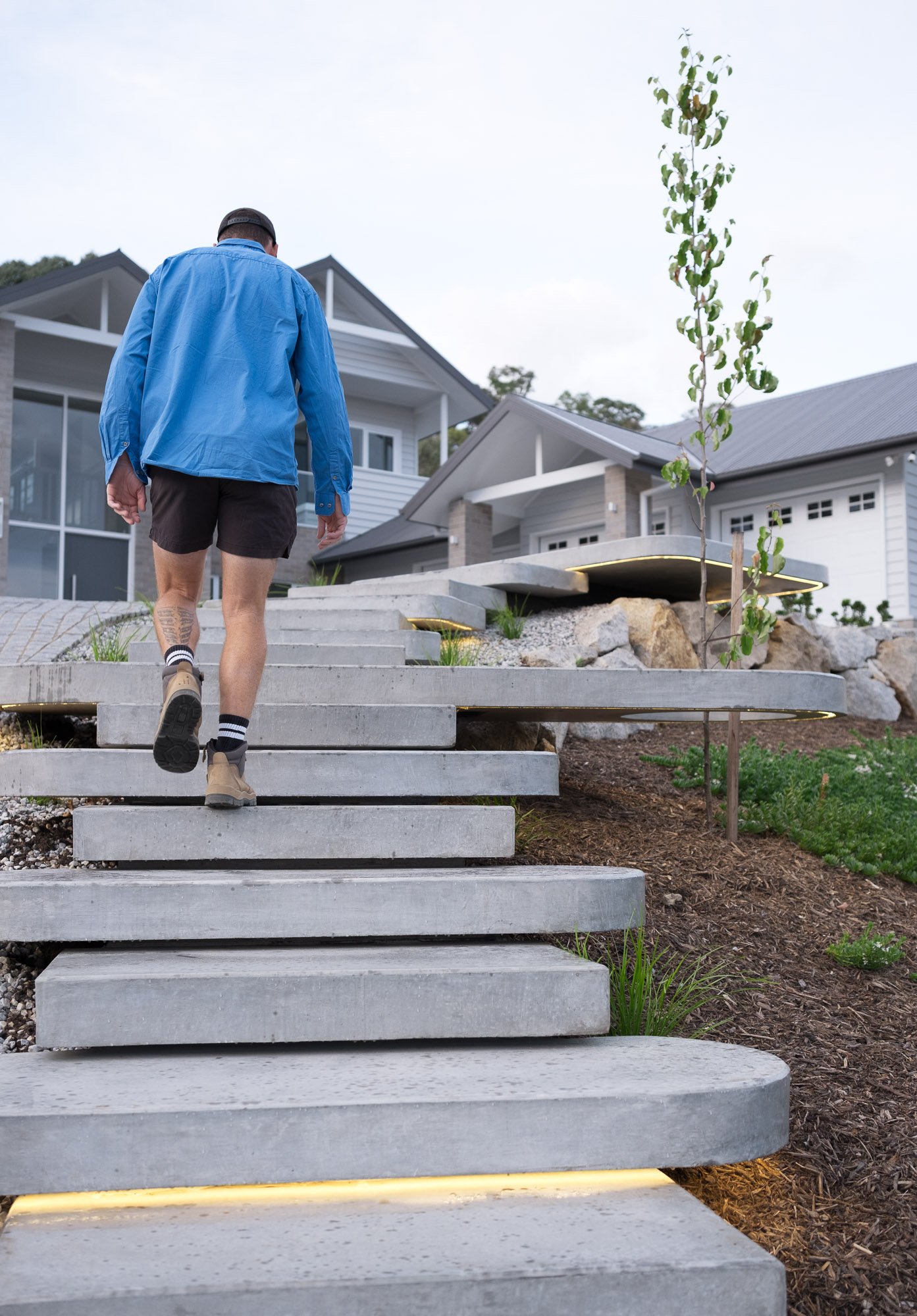 A person dressed in a blue jacket, black shorts, and athletic shoes walking up concrete stairs outside a modern house with large windows and a metal roof, with a young tree and landscaping on the side.