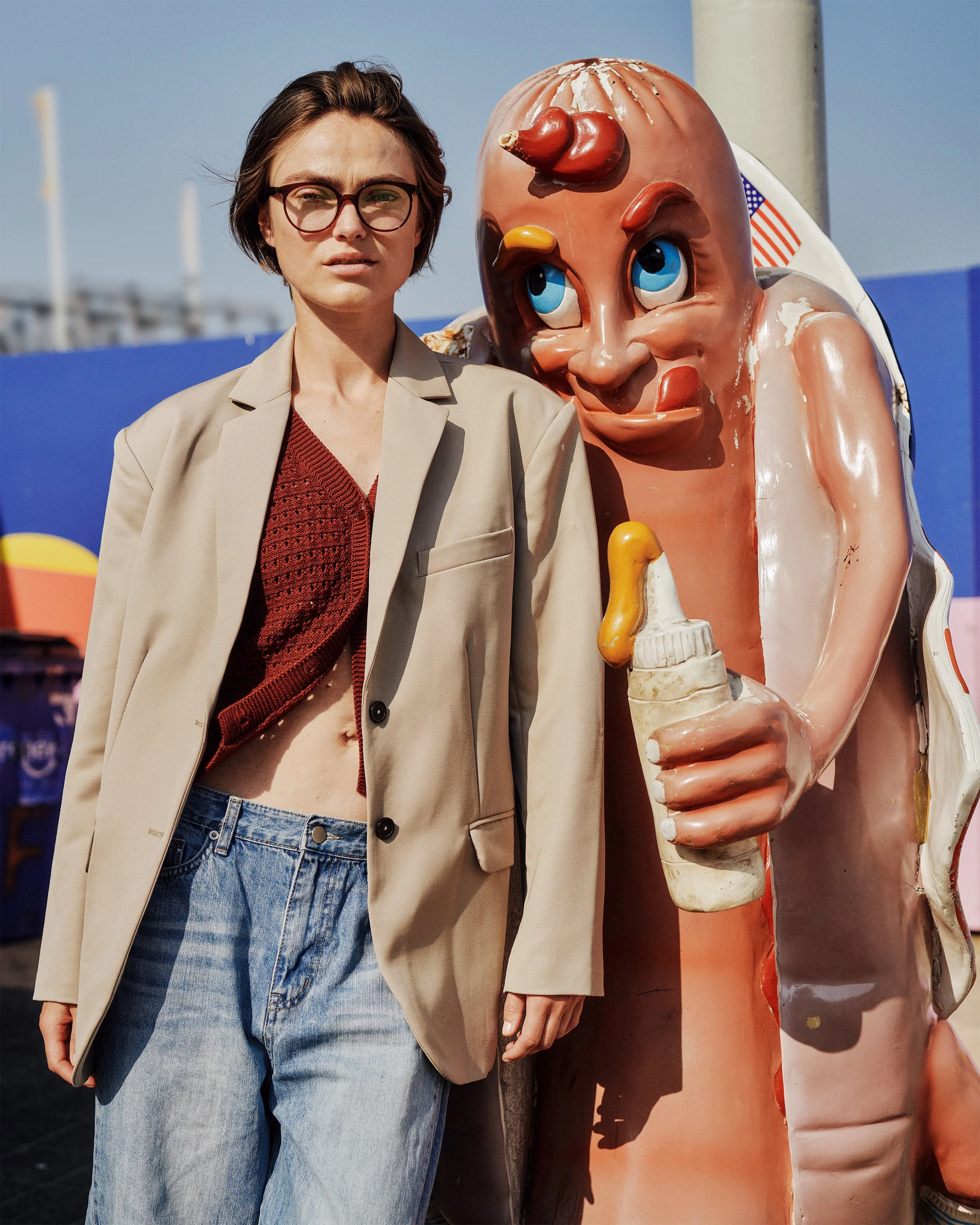 Person standing beside a hot dog mascot statue, wearing a suit jacket and jeans, with a blue sky in the background.