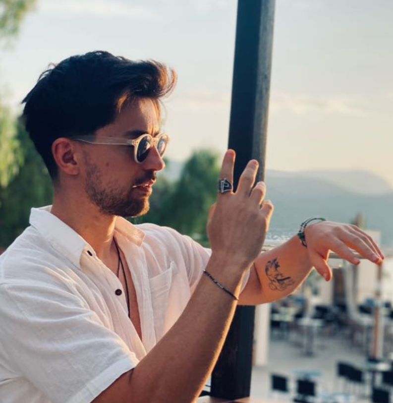 A person wearing sunglasses and a white shirt gesturing with their hands outdoors, with a view of trees and mountains in the background.