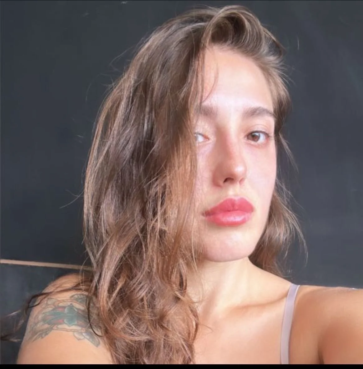 Close-up of a person with long hair, wearing minimal makeup, against a dark background. The person's shoulder is visible, featuring a tattoo.
