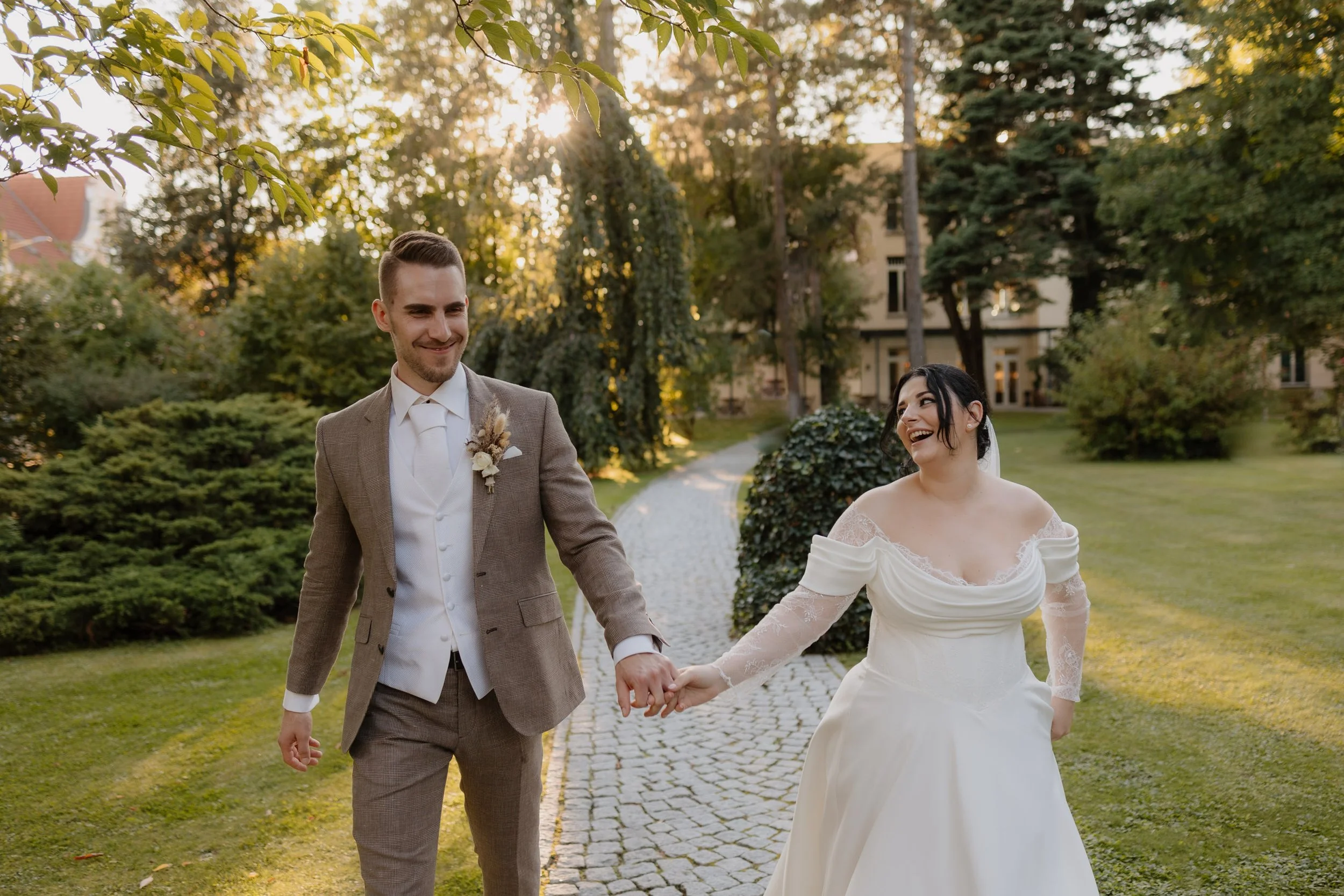Ein lachendes Brautpaar beim Spaziergang im Garten bei Sonnenuntergang, die Frau trägt ein weißes Hochzeitskleid und der Mann trägt einen braunen Anzug.
