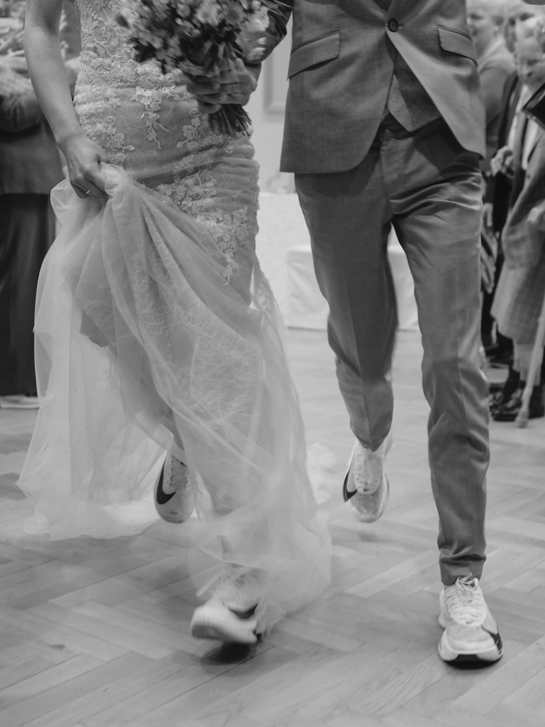 Ein Brautpaar läuft lachend durch eine versammelte Gruppe von Gästen bei einer Hochzeit. Beide sind glücklich und die Gäste applaudieren und feiern sie.