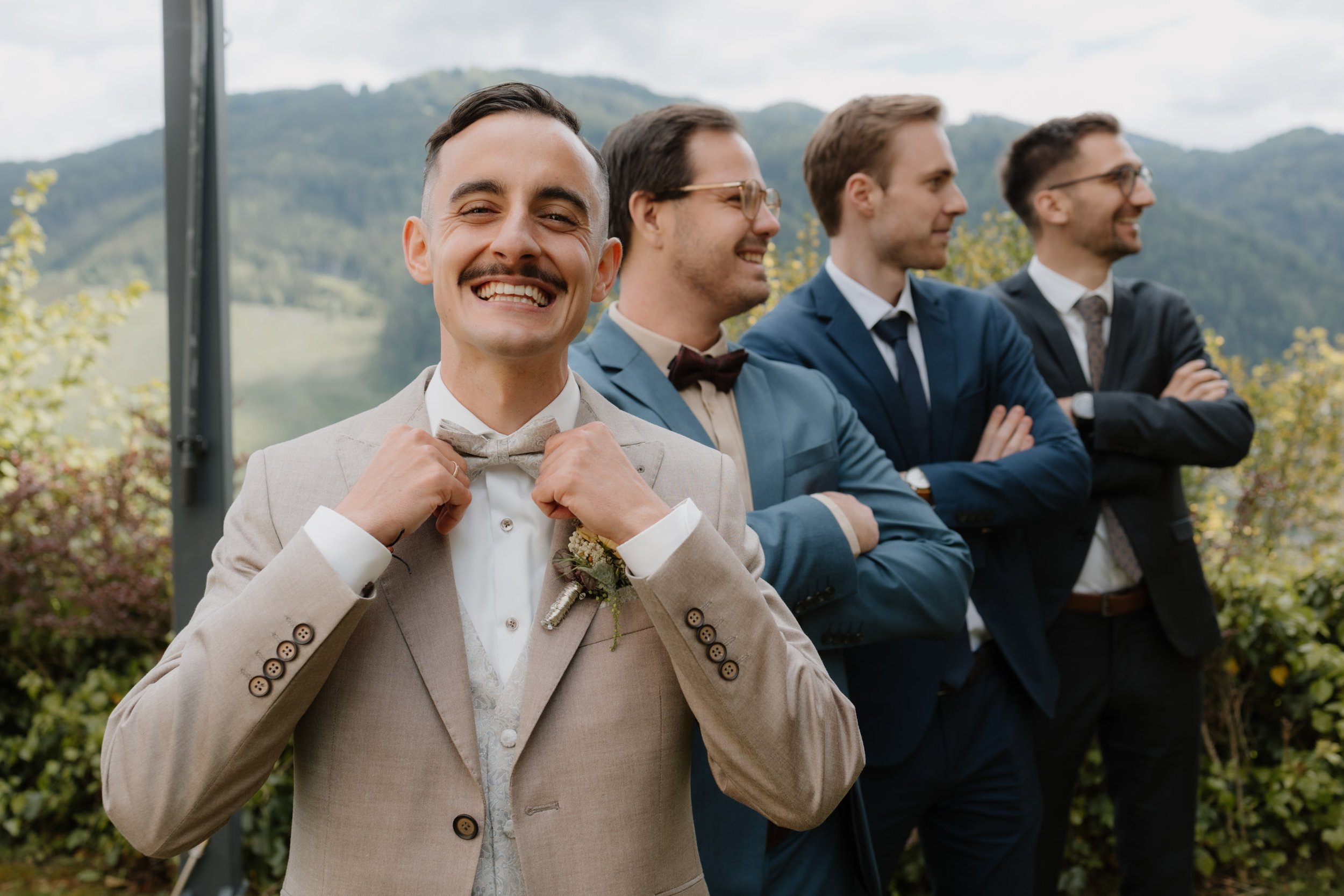 Junggesellen gruppe Foto. Junge Männer in Anzügen bei einem outdoor Event, mit Bergen im Hintergrund, lachend und lässig.