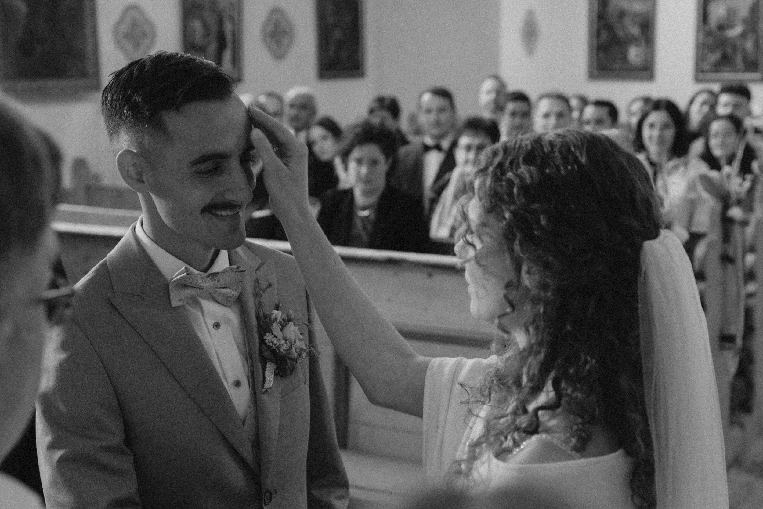 Ein Mann in einem Anzug und eine Frau in einem weißen Kleid bei einer Hochzeit, die eine emotionale Geste zeigt, während die Zuschauer im Hintergrund zusehen.