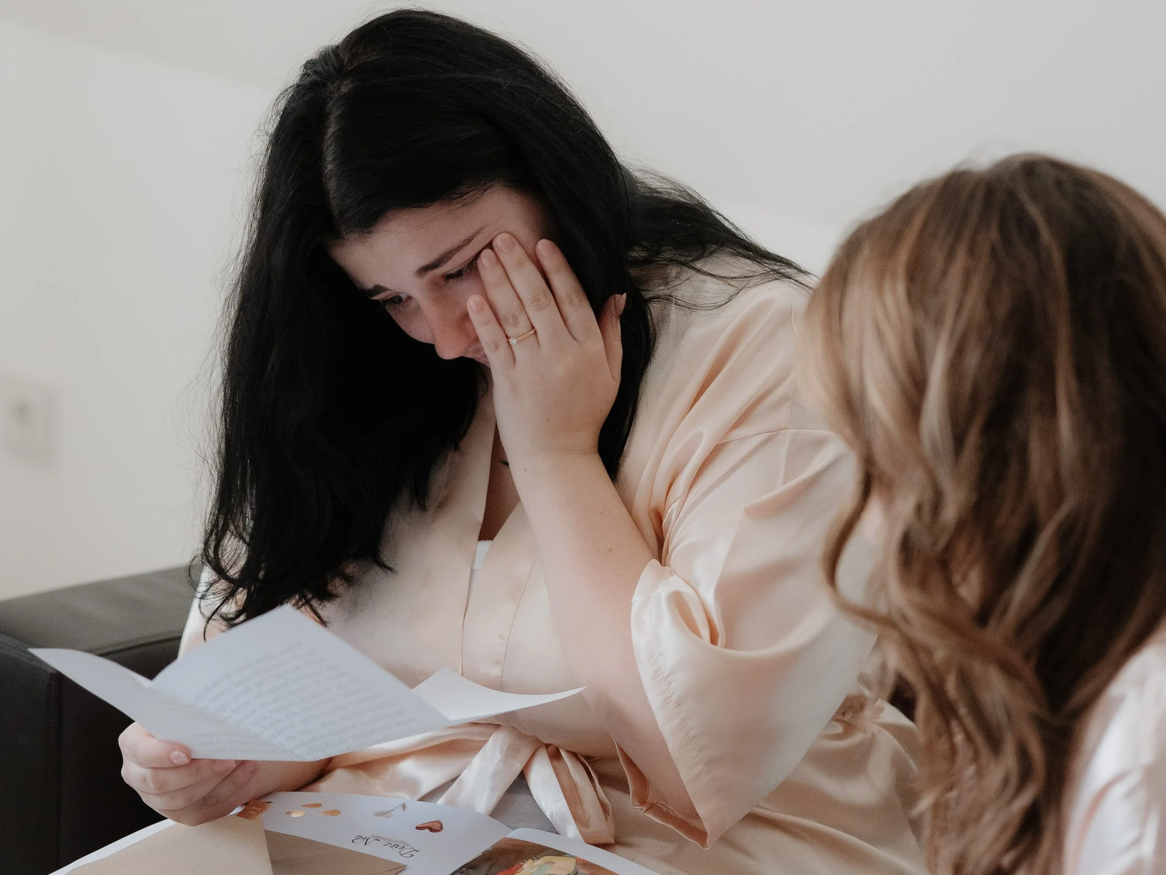 Eine Frau mit langen schwarzen Haaren liest einen Brief mit einer Hand. Sie trägt ein hellrosa Oberteil und sitzt an einem Tisch, die Augen sind überrascht oder emotional.