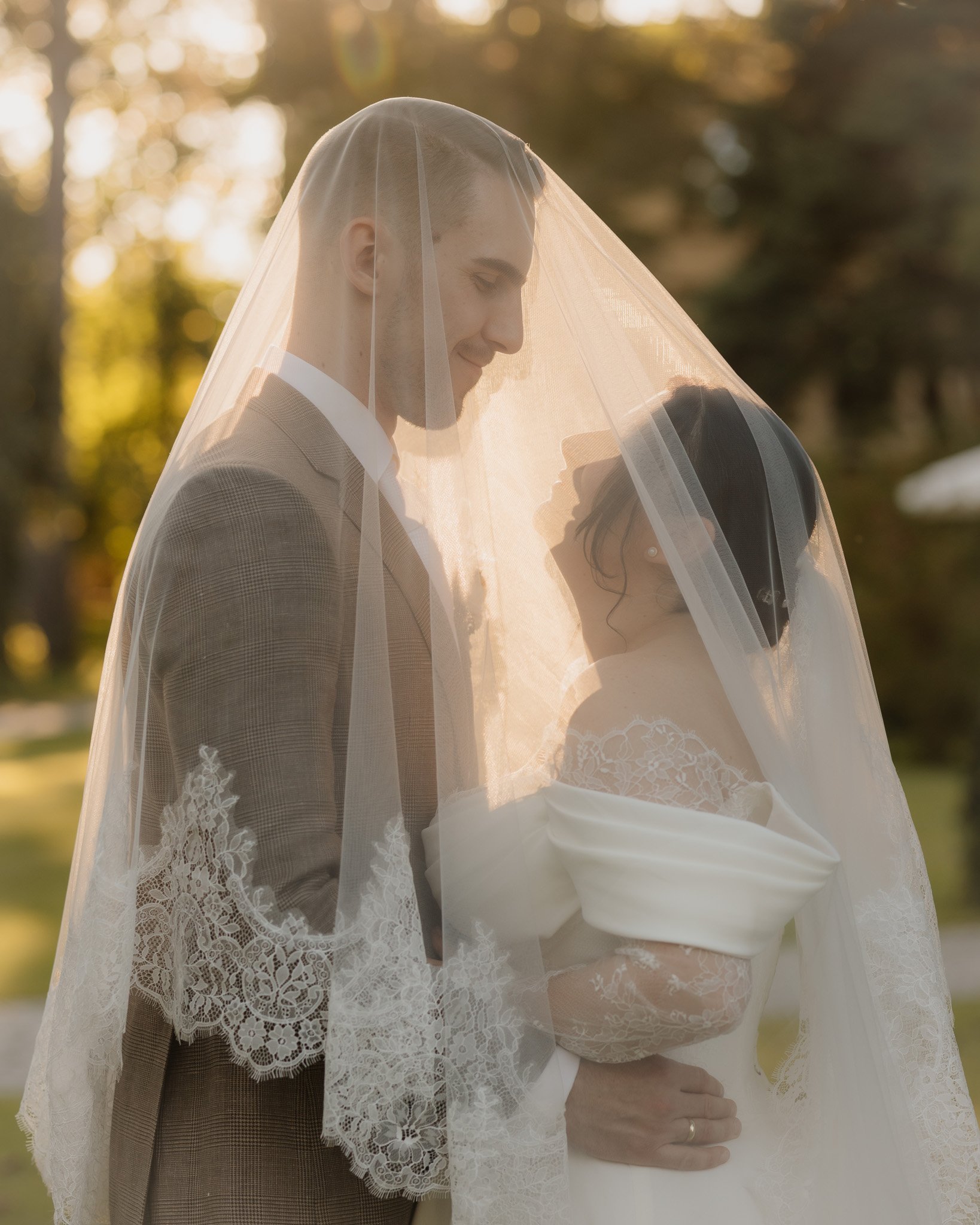 Ein Paar bei einer Hochzeit, die Braut trägt ein weißes Hochzeitskleid mit Spitzenarmringen, der Bräutigam trägt einen hellbraunen Anzug und eine weiße Krawatte. Sie stehen im Freien bei Sonnenuntergang, umgeben von Bäumen.