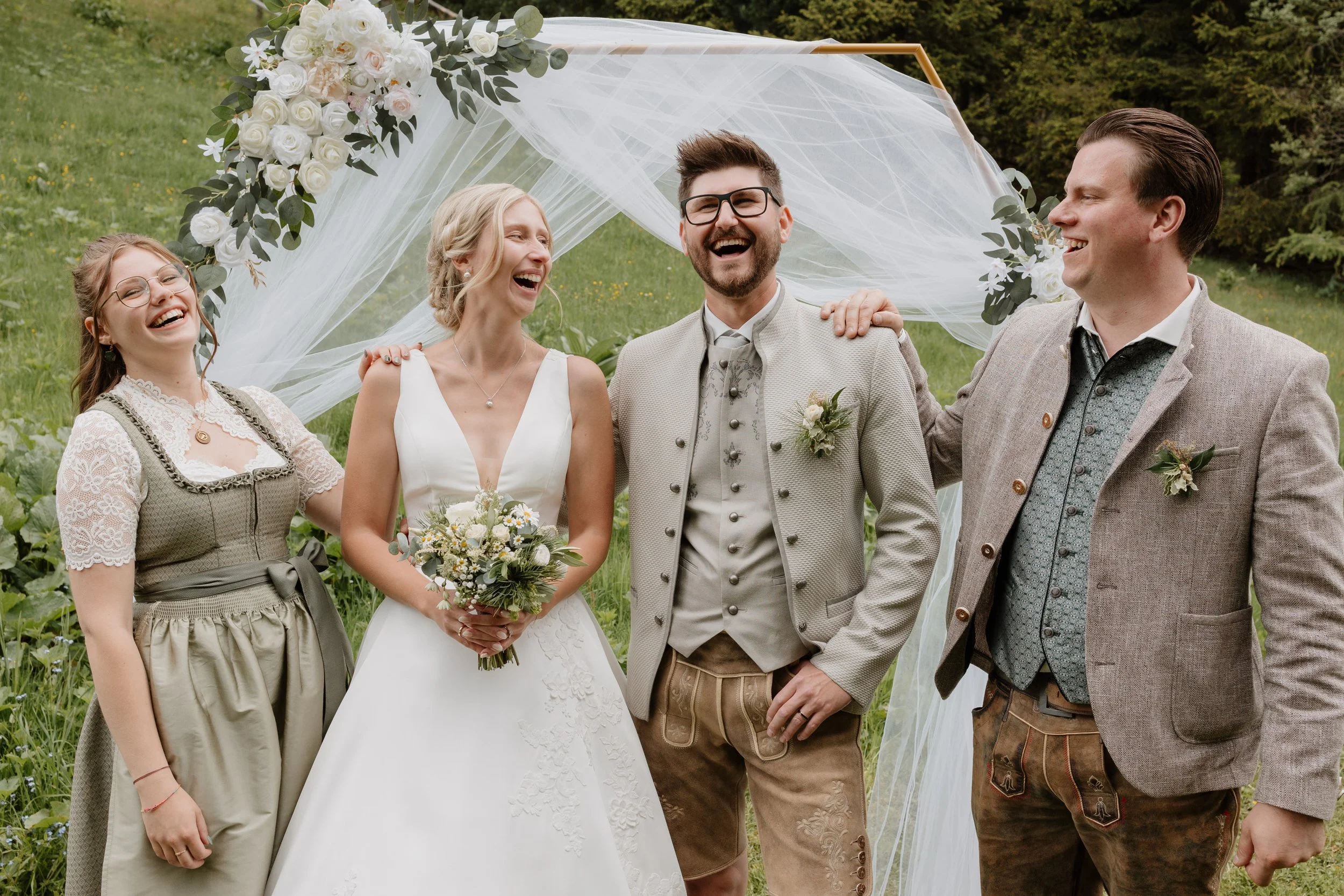 Eine fröhliche Gruppe von vier Menschen bei einer Hochzeit im Freien, mit einem blumenverzierten Bogen im Hintergrund.