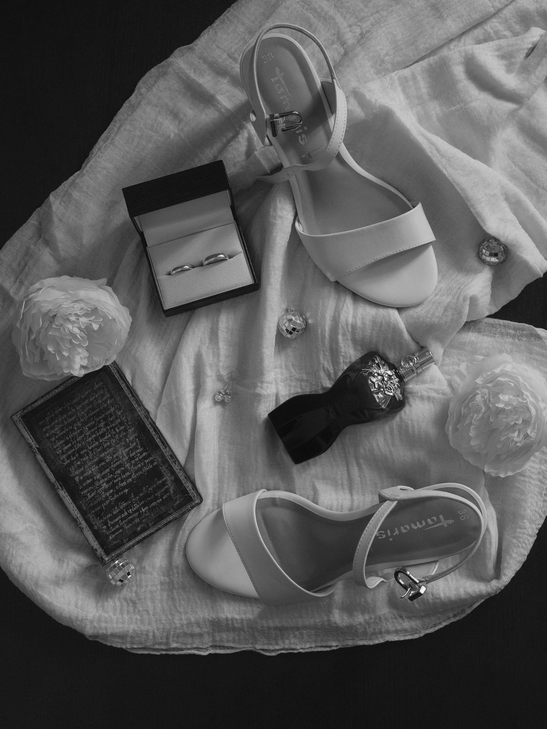 Schwarz-weiß-Fotografie von mehreren Hochzeits-Accessoires auf einem weißen Tuch, darunter weiße Hochzeitsschuhe, ein Polaroid-Kästchen mit Eheringen, eine blumenartige Dekoration, ein Parfümflakon, eine kleine Tafel mit handgeschriebenem Text, und e