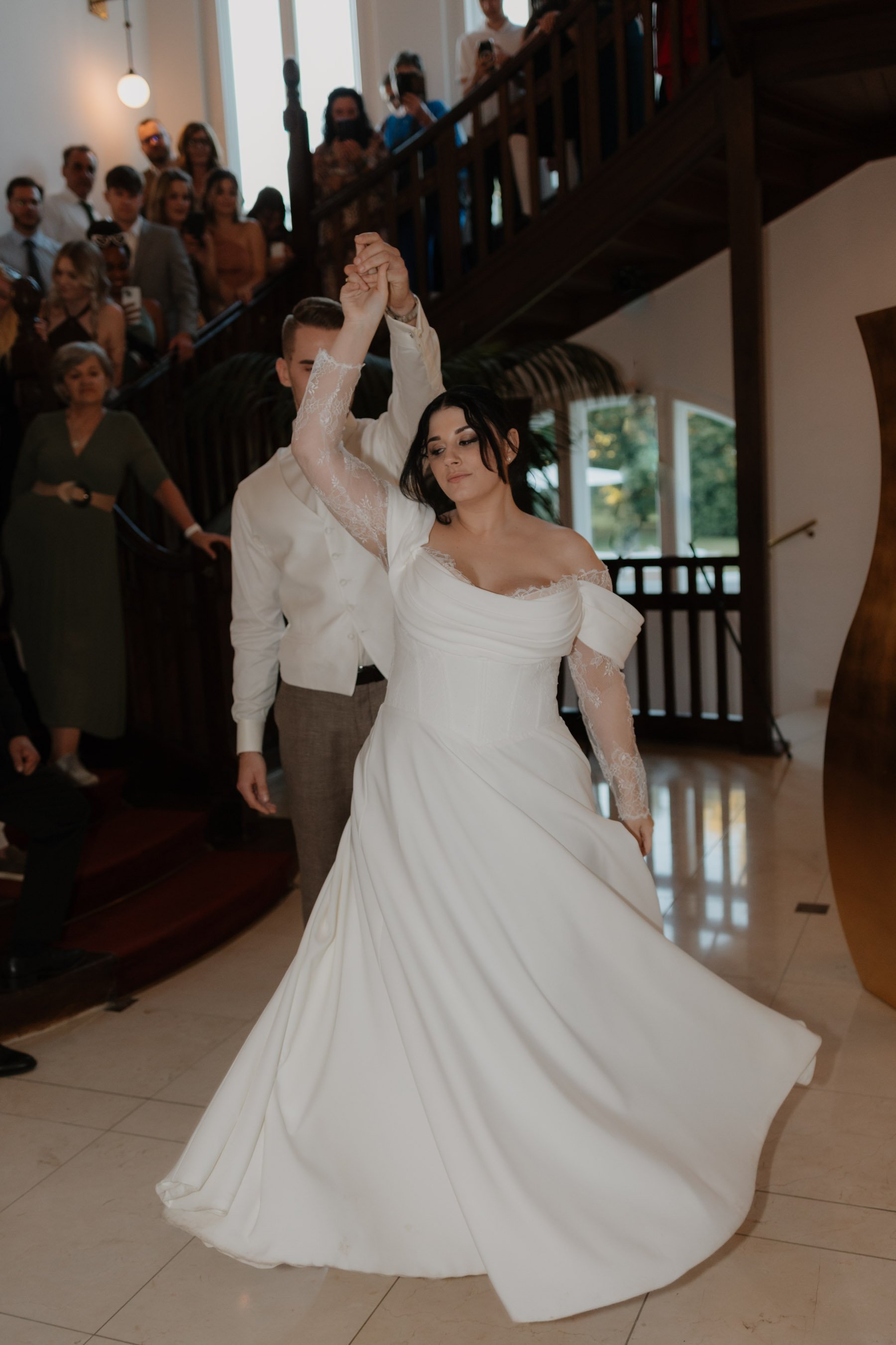 Ein Brautpaar tanzt bei einer Hochzeit, die Braut trägt ein weißes Kleid, der Bräutigam einen Anzug, im Hintergrund sehen wir Gäste und eine Treppe.