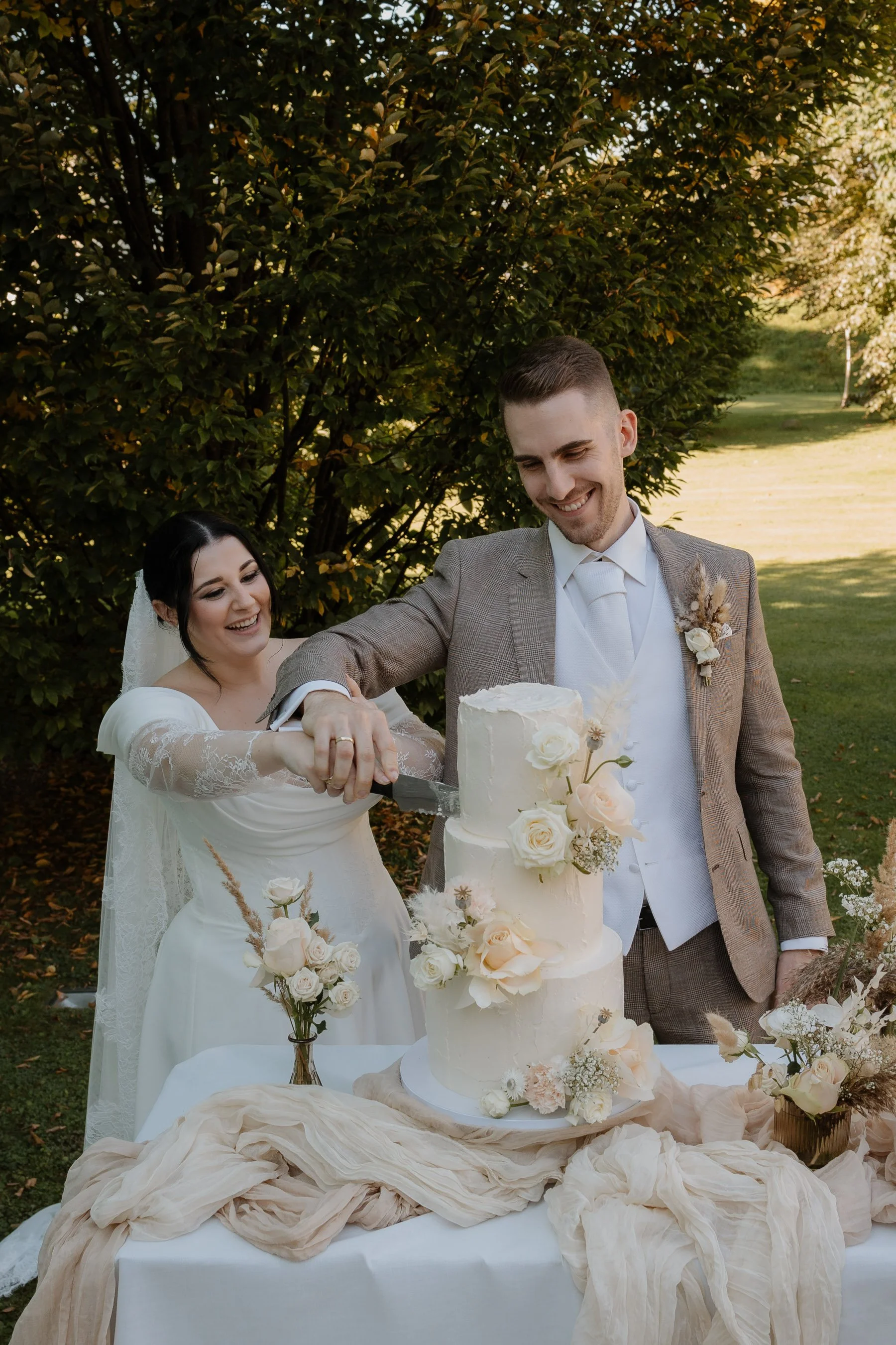 Ein Hochzeitspaar schneidet eine Hochzeitstorte im Freien, umgeben von Blumen und Bäumen, bei Sonnenuntergang.