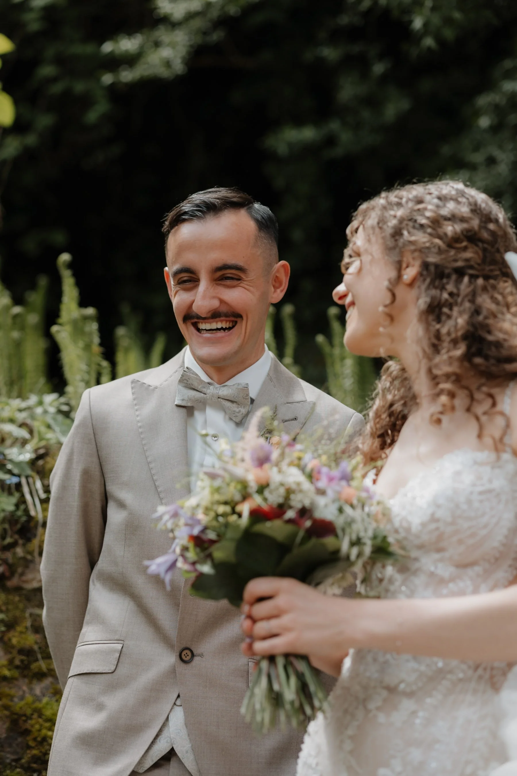Ein lachendes Ehepaar bei ihrer Hochzeit, der Mann trägt einen hellgrauen Anzug mit Fliege, die Frau trägt ein weißes Hochzeitskleid und hält einen Blumenstrauß, im Hintergrund grüne Pflanzen und Bäume.
