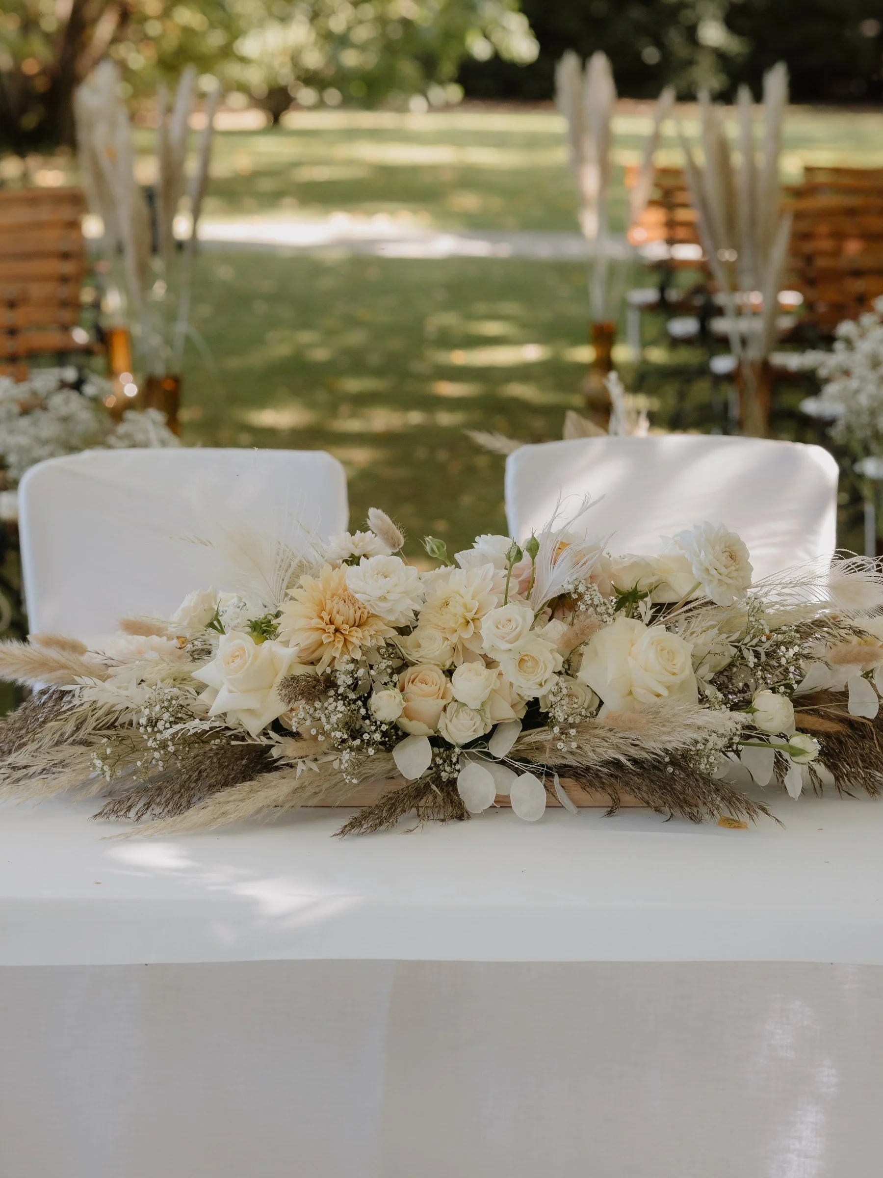 Detailaufnahmen Hochzeit. Blumenarrangement auf einem Tisch, im Hintergrund eine outdoor Zeremonie mit aufgestellten Stühlen.