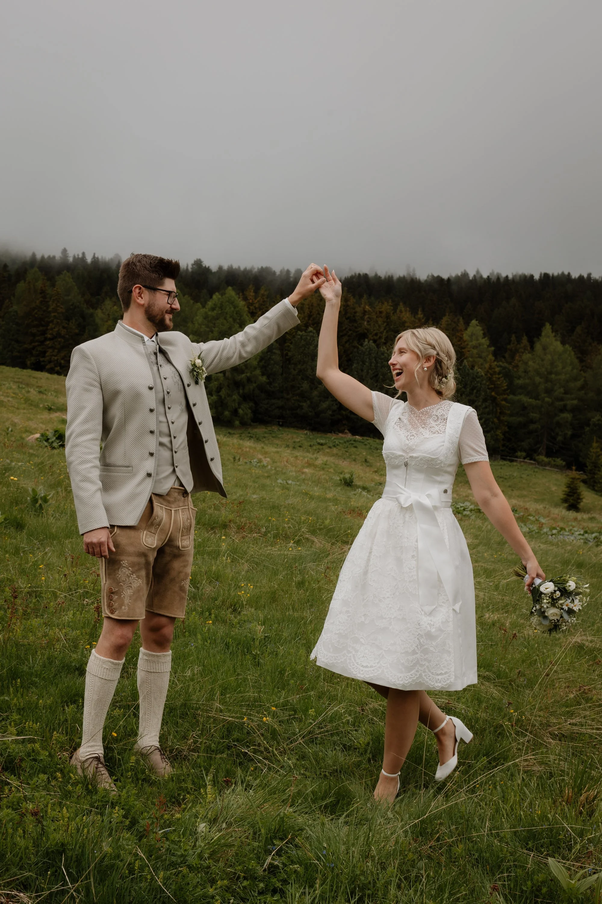 Ein Mann und eine Frau, beide in Trachtenkleidung, tanzen lachend auf einer grünen Wiese mit einem Wald im Hintergrund, während sie sich an den Händen halten.