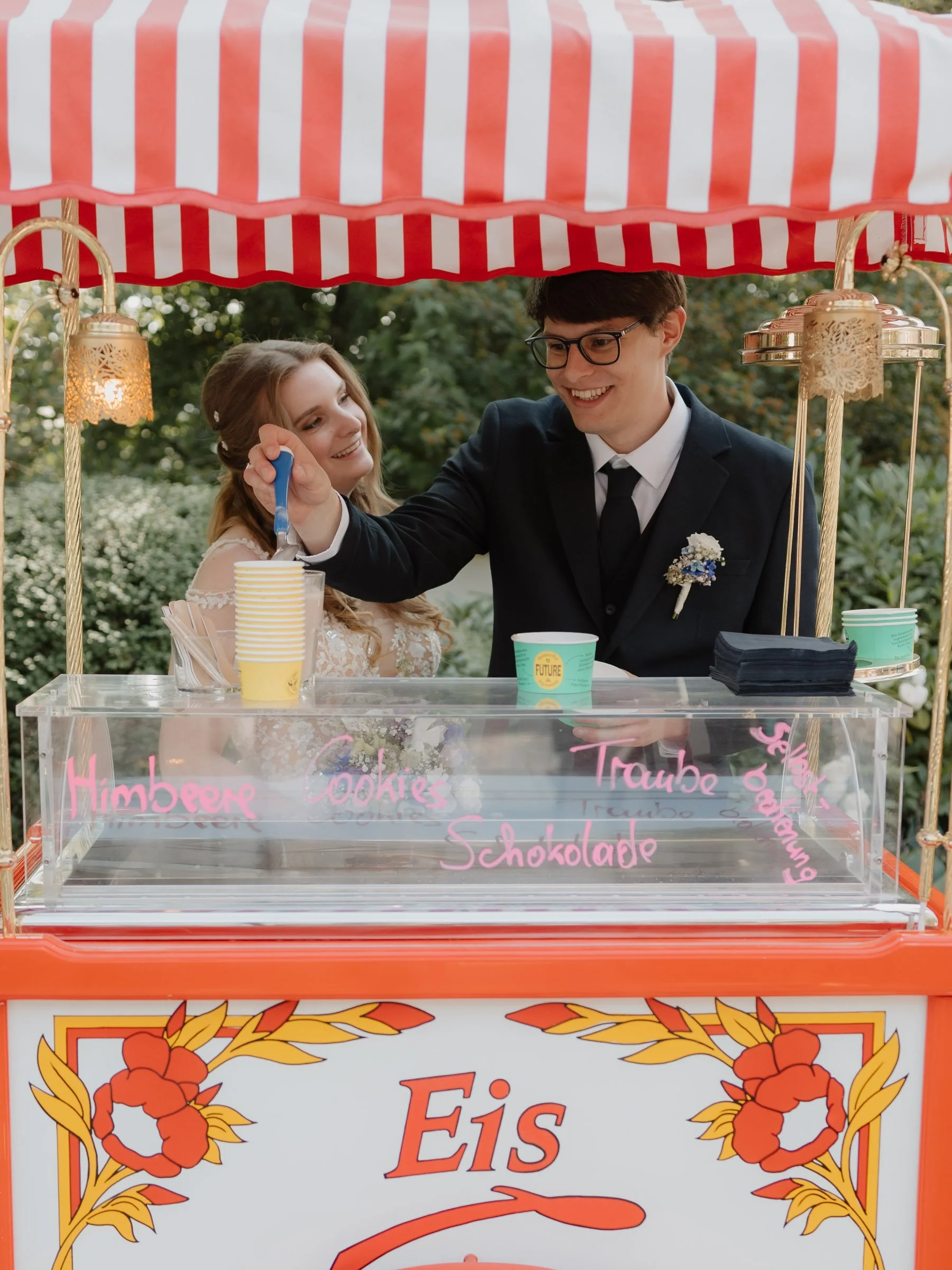 Ein Eiswagen mit einem rot-weißen Dach, auf dem zwei Menschen stehen, die Eis in Becher servieren. Der Wagen ist mit bunten Farben und Dekorationen verziert, und es sind Schilder mit Eissorten wie Himbeere, Cookies, Traube, Schokolade sichtbar.