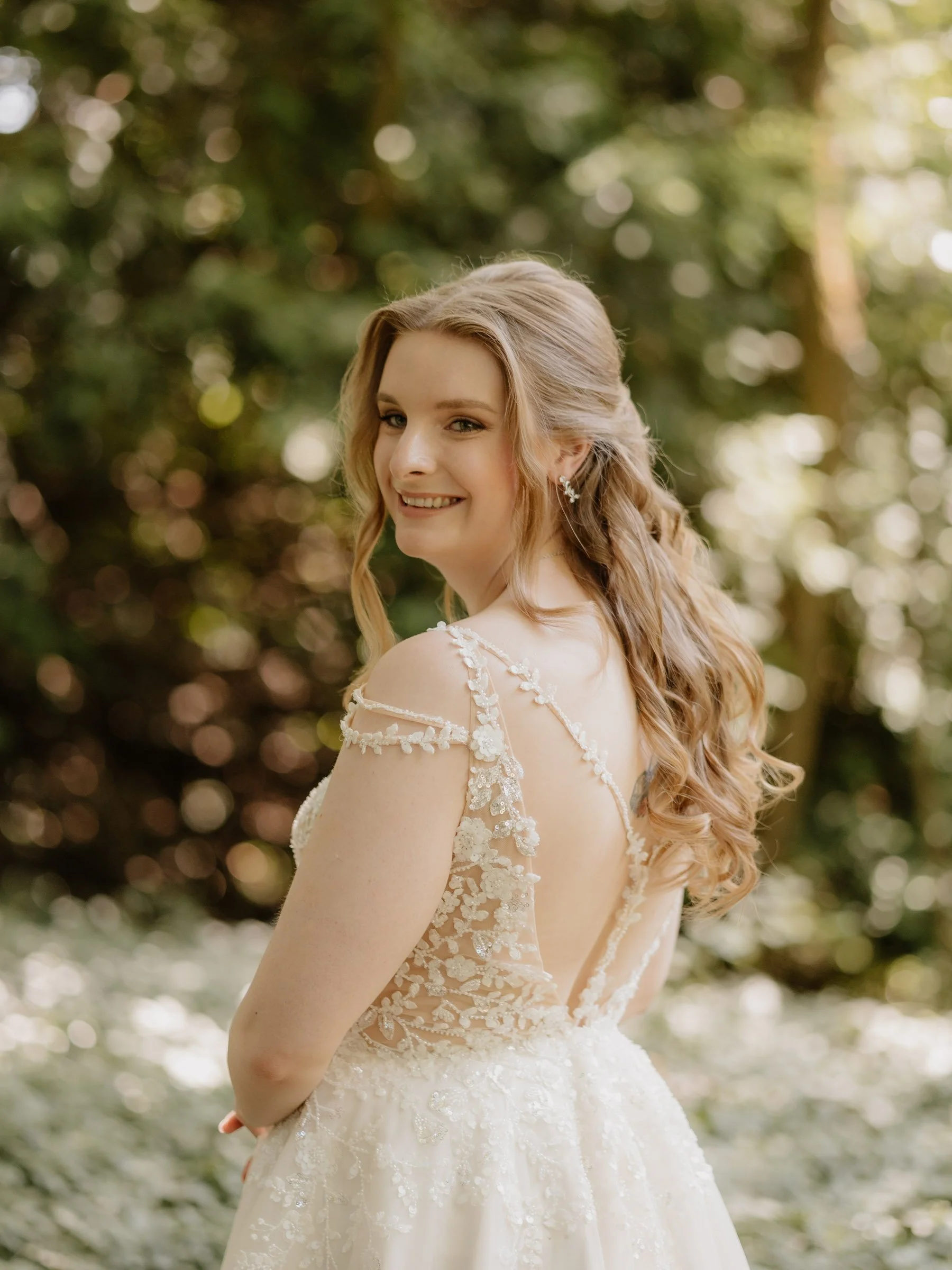 Eine lachende Braut in einem weißen Brautkleid, mit lockigem Haar, vor einem unscharfen grünen Baumhintergrund.