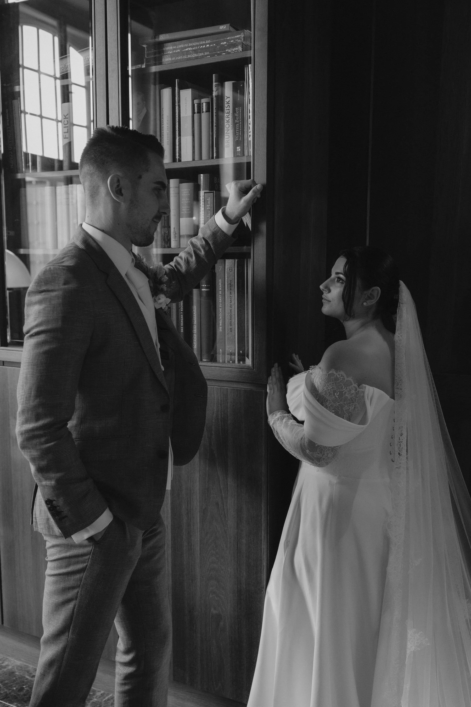 Ein Mann in einem Anzug steht vor einem Bücherregal, während eine Frau in einem Hochzeitskleid neben ihm steht, beide scheinen in einem emotionalen Moment während einer Hochzeit zu sein.