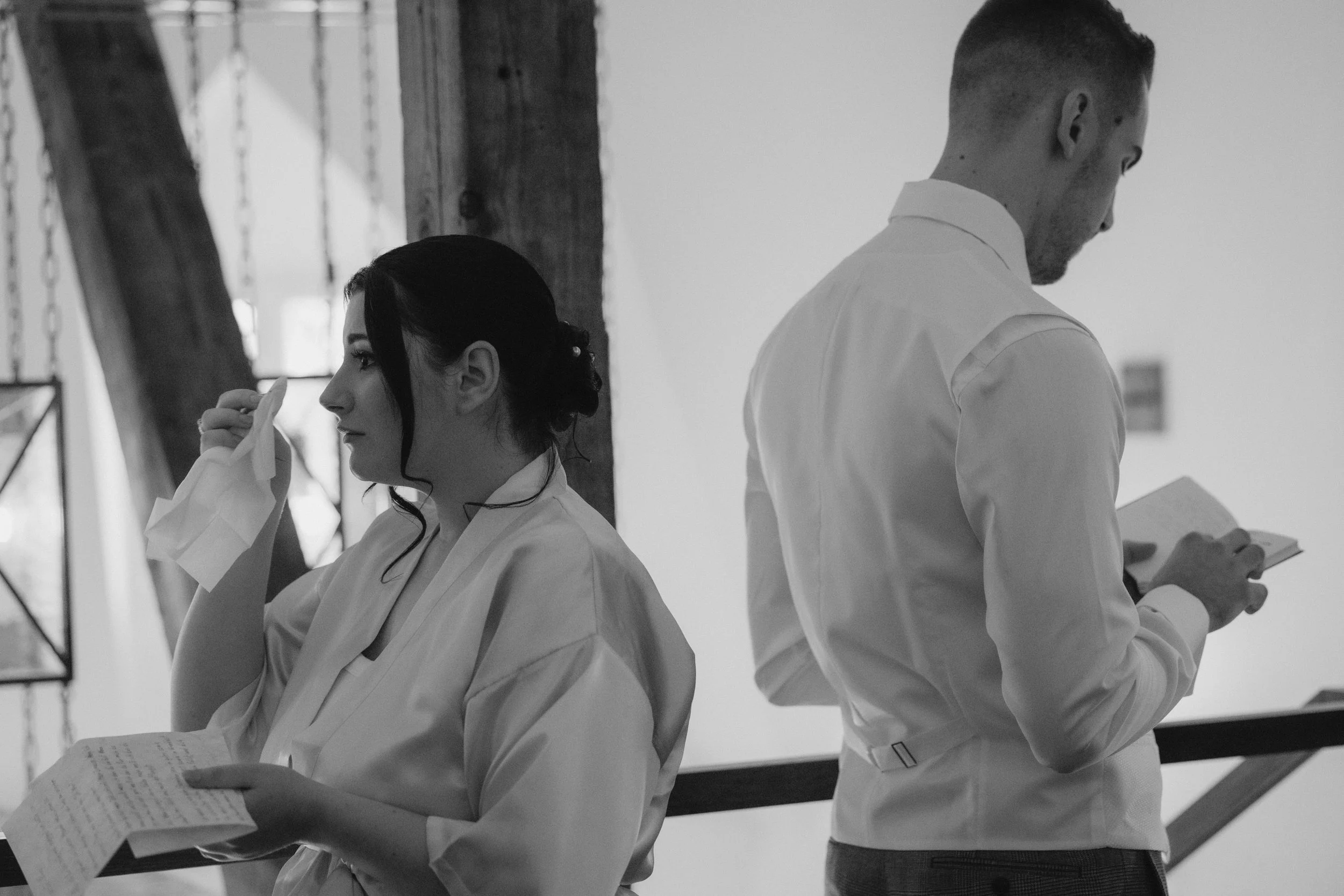 Secret Vows, Ehegelübte Fotos. Ein Mann und eine Frau stehen nebeneinander und lesen in Papieren, während sie in einem Raum mit Holzbalken und einer Kletterwand im Hintergrund sind.
