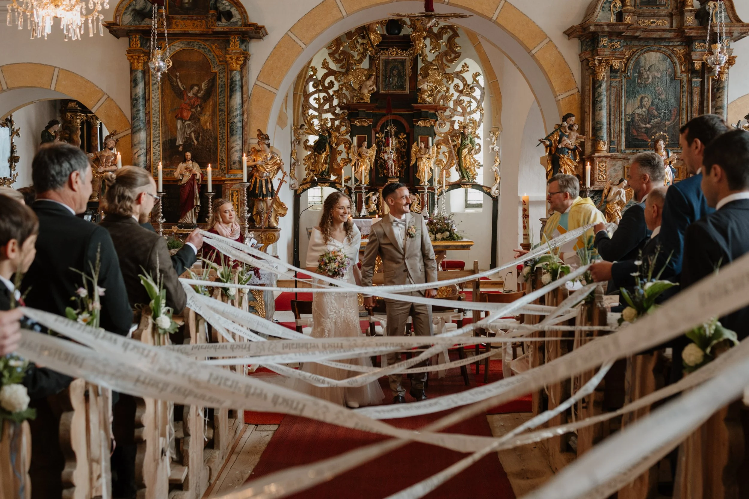 Ein Brautpaar steht in einer Kirche während ihrer Hochzeitszeremonie, umgeben von Freunden und Familienmitgliedern. Im Hintergrund sind barocke Kirchenaltäre mit goldverzierten Skulpturen und Gemälden. Die Kirche ist reich verziert mit Kerzen, Blumen