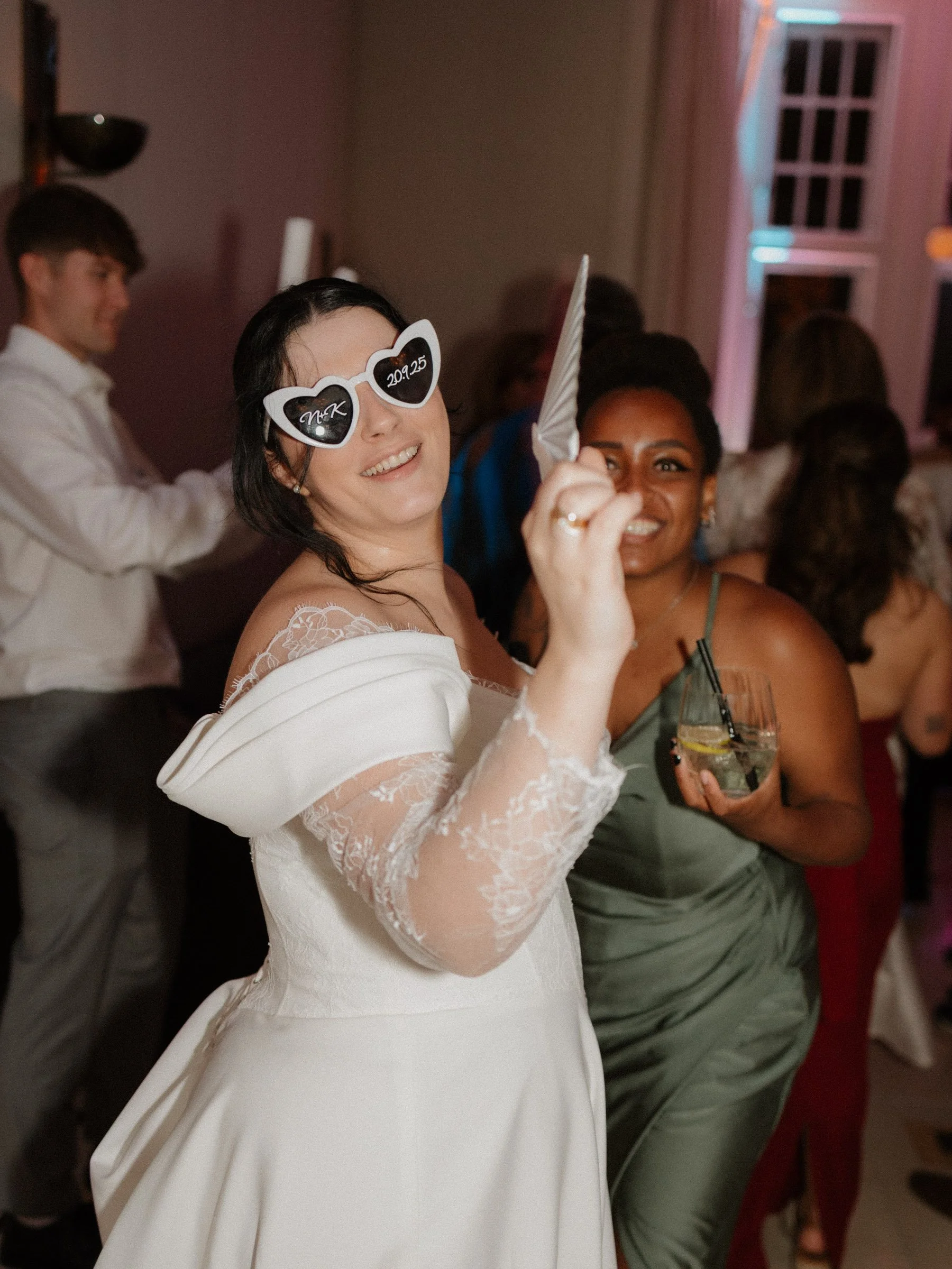 Hochzeitsparty Fotos. Eine lachende Braut mit dunklen Haaren trägt Herzbrille. Sie trägt ein weißes Kleid mit Spitzenärmeln und hält einen Fächer in der Hand. 