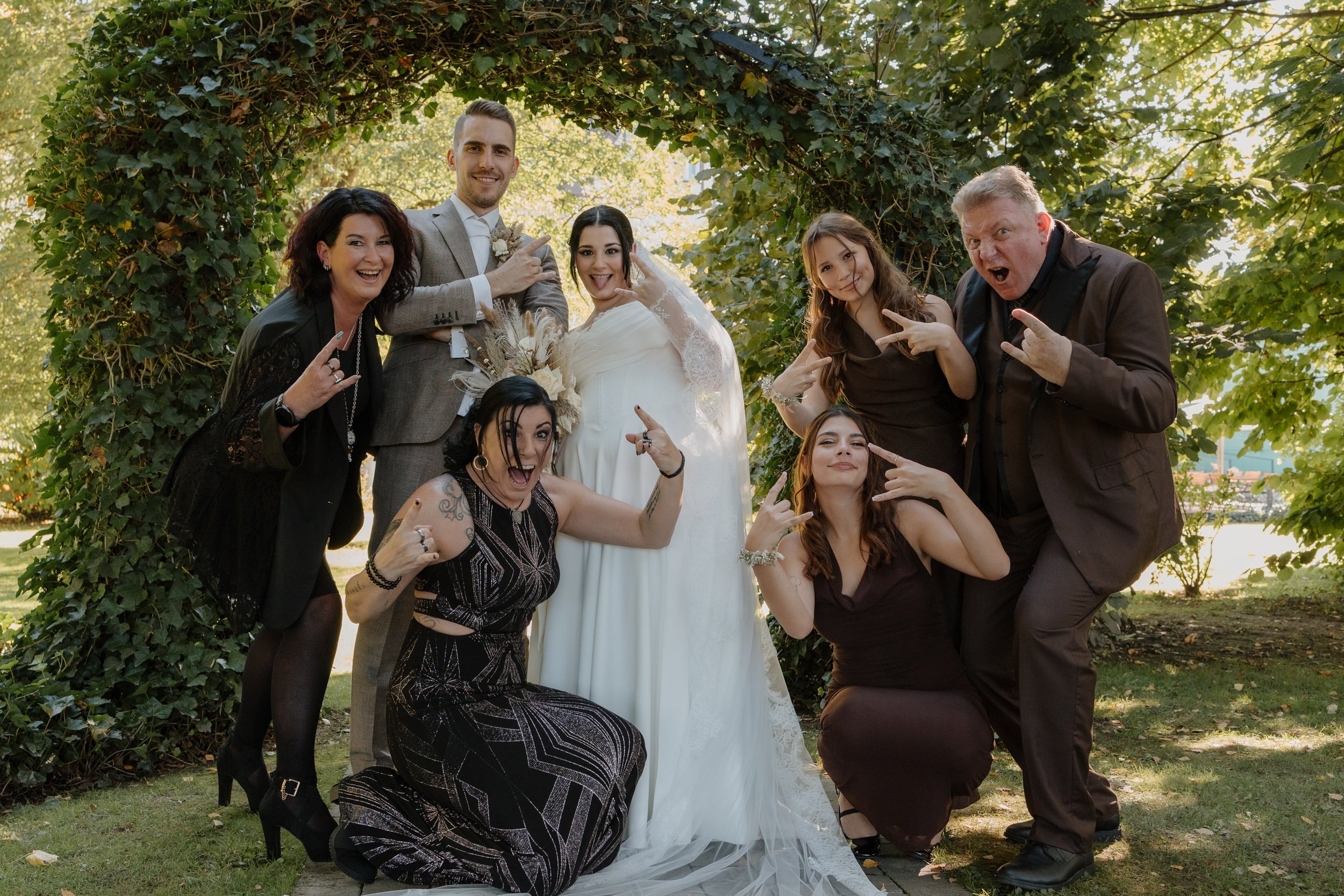 Eine Gruppe von sieben Menschen, darunter ein Brautpaar, posiert fröhlich im Freien unter einem grünen Baumbogen für ein Foto, alle machen Rock- und Roll-Gesten, die Stimmung ist ausgelassen und feierlich.